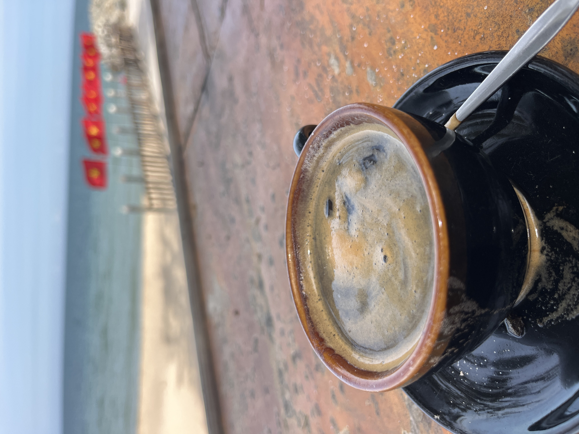 A close-up shot features a steaming cup of black coffee on a dark saucer, placed on a textured, orange-brown surface, possibly a table. A silver spoon rests on the saucer, partially submerged in the liquid. In the blurred background, a beach stretches towards the ocean, with a line of red flags with yellow stars, likely Vietnamese flags, visible along the shore. The sky above is a pale blue, meeting the calmer, greenish-blue of the sea. The scene suggests a relaxed setting, likely a cafe or restaurant overlooking the beach in Mũi Né, Vietnam.