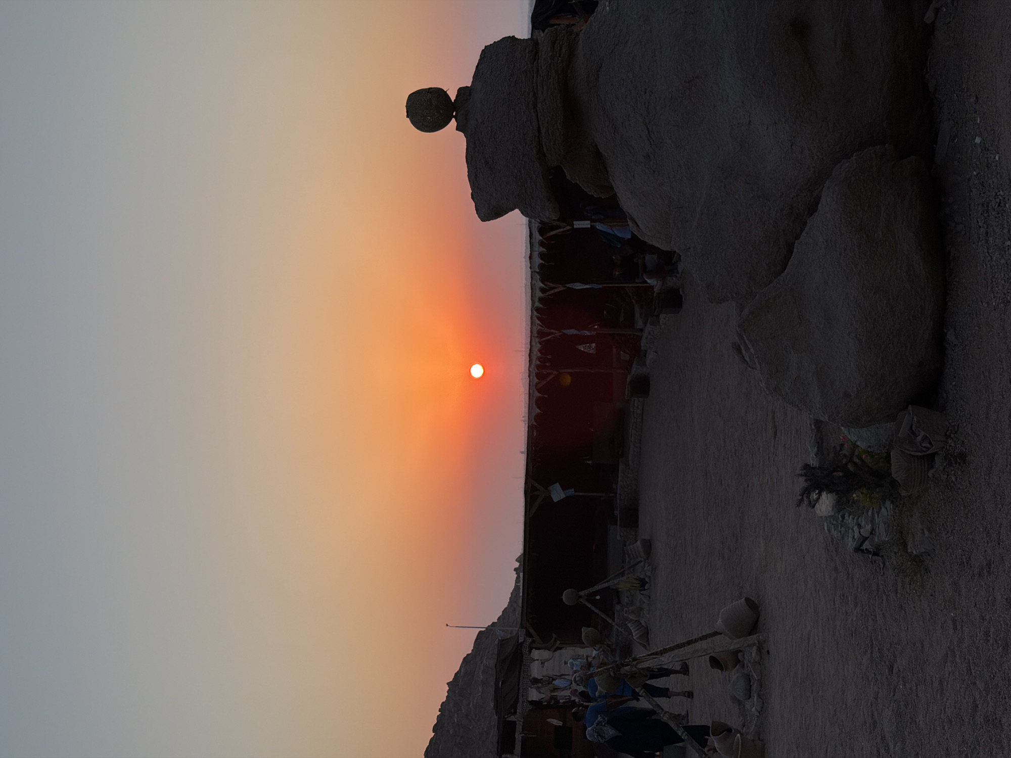 The photograph captures a sunset scene at a desert location, likely in Sharm El Sheikh, Egypt, given the characteristic arid landscape and warm, vibrant sky. The sun, a bright orb of orange, is low on the horizon, casting a warm glow across the scene and painting the sky in hues of orange, pink, and pale blue.
In the foreground, large, rough-hewn rocks dominate the right side of the frame. One of these rocks has an unusual, almost sculpted appearance, topped with a smaller, rounded stone, resembling a primitive monument. The ground is composed of sand and small pebbles, with scattered pottery and what appear to be clay amphorae positioned around a wooden tripod structure. A small, sparse plant adds a touch of green to the otherwise earthy tones.
In the mid-ground, a collection of tents or market stalls with canvas coverings are visible, suggesting a temporary settlement or marketplace. Several individuals are present, some walking in the sandy area, others possibly engaged in activities within or around the stalls. One person in the left mid-ground wears a headscarf, while another in the foreground appears to be walking away from the camera. In the distance, the silhouette of a rocky hill or mountain range rises against the sunset sky. The overall atmosphere is one of serene desert beauty, tinged with the subtle activity of human presence.