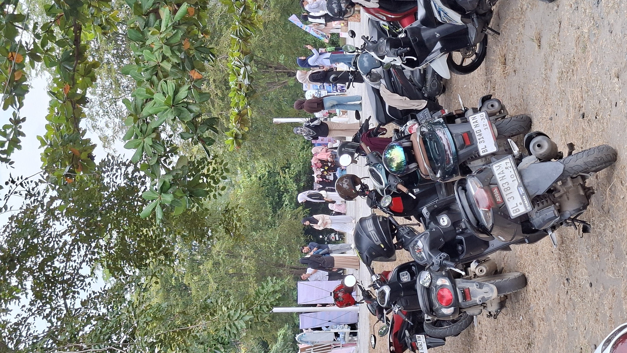 A group of motorcycles is parked on a sandy, dry ground area. In the background, several people are standing and walking around what appears to be a market or event with stalls and displays. The environment is outdoors with trees and foliage visible, suggesting a park or open public space. The scene captures a moment of everyday life in Lhokseumawe, Indonesia, likely a marketplace or community gathering.