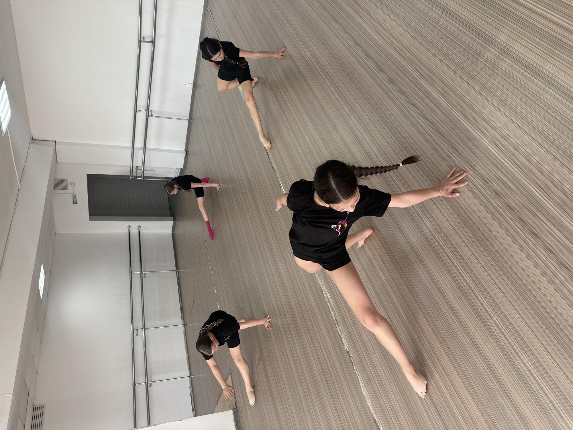 The image shows a dance studio in Votkinsk, Russia. Four young women in black t-shirts and black shorts are practicing a modern dance routine. They are barefoot, and their bodies are positioned in various poses, suggesting a flowing, synchronized movement. The studio has a light-colored, wood-patterned floor and white walls with a barre system along one side. A mirror is visible at the edge of the image, reflecting the dancers and the studio environment. The lighting is bright and even.