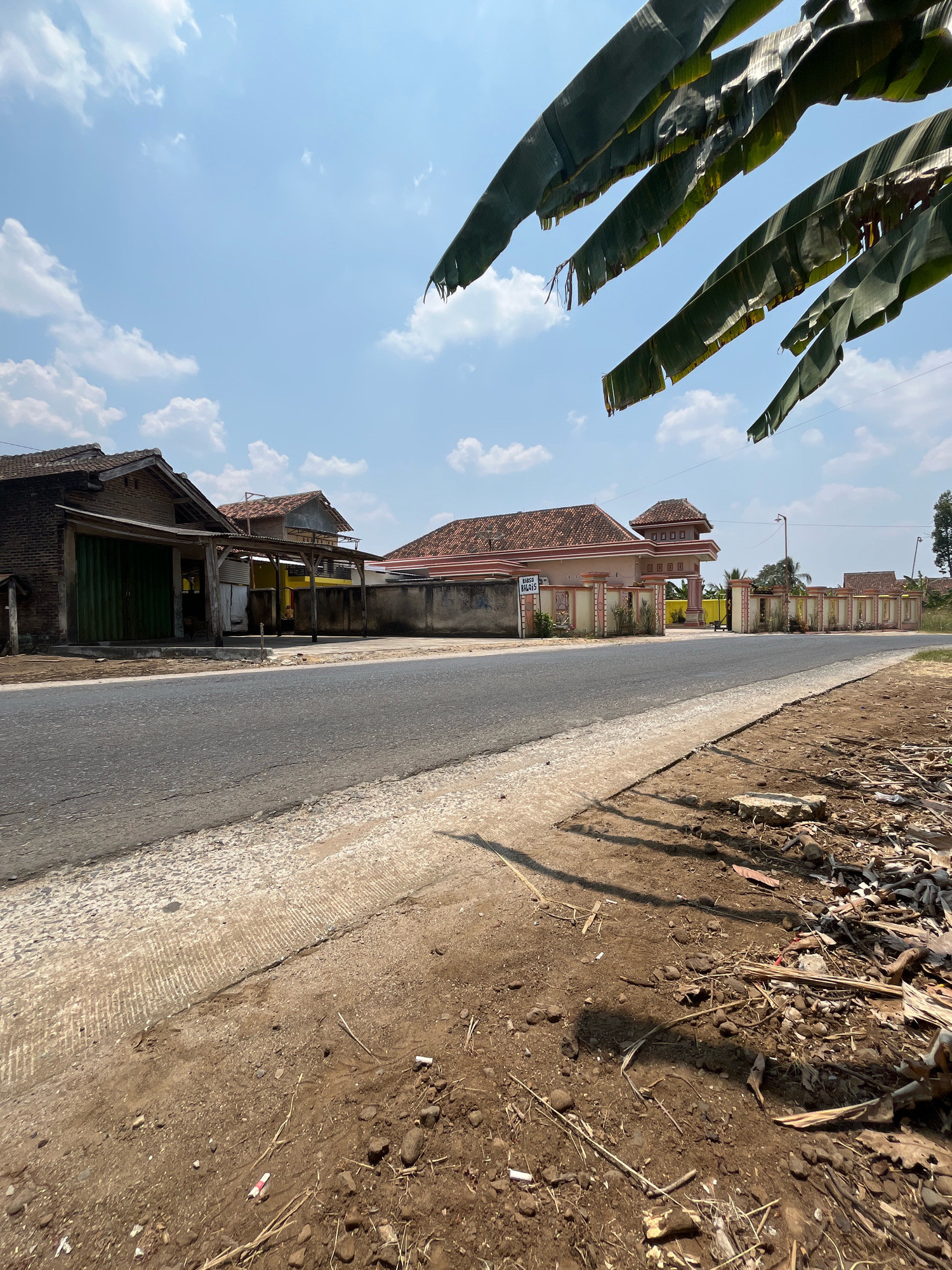 A paved road runs through a rural area in Penantian, Indonesia. The road is bordered on the left by a rough, unpaved shoulder with debris, and on the right by a manicured yard with a fence and vegetation. Several buildings are visible along the road, including a dark brick building with a green metal roll-up door and a lighter-colored building with a tiled roof and an open-air porch. Further down the road, a larger building with a decorative gate and a tower-like structure is visible. The sky is blue with scattered white clouds, and the image is framed by large green leaves in the foreground, suggesting they are from a banana tree. The scene appears to be captured during daylight, likely midday, given the bright sunlight and distinct shadows.