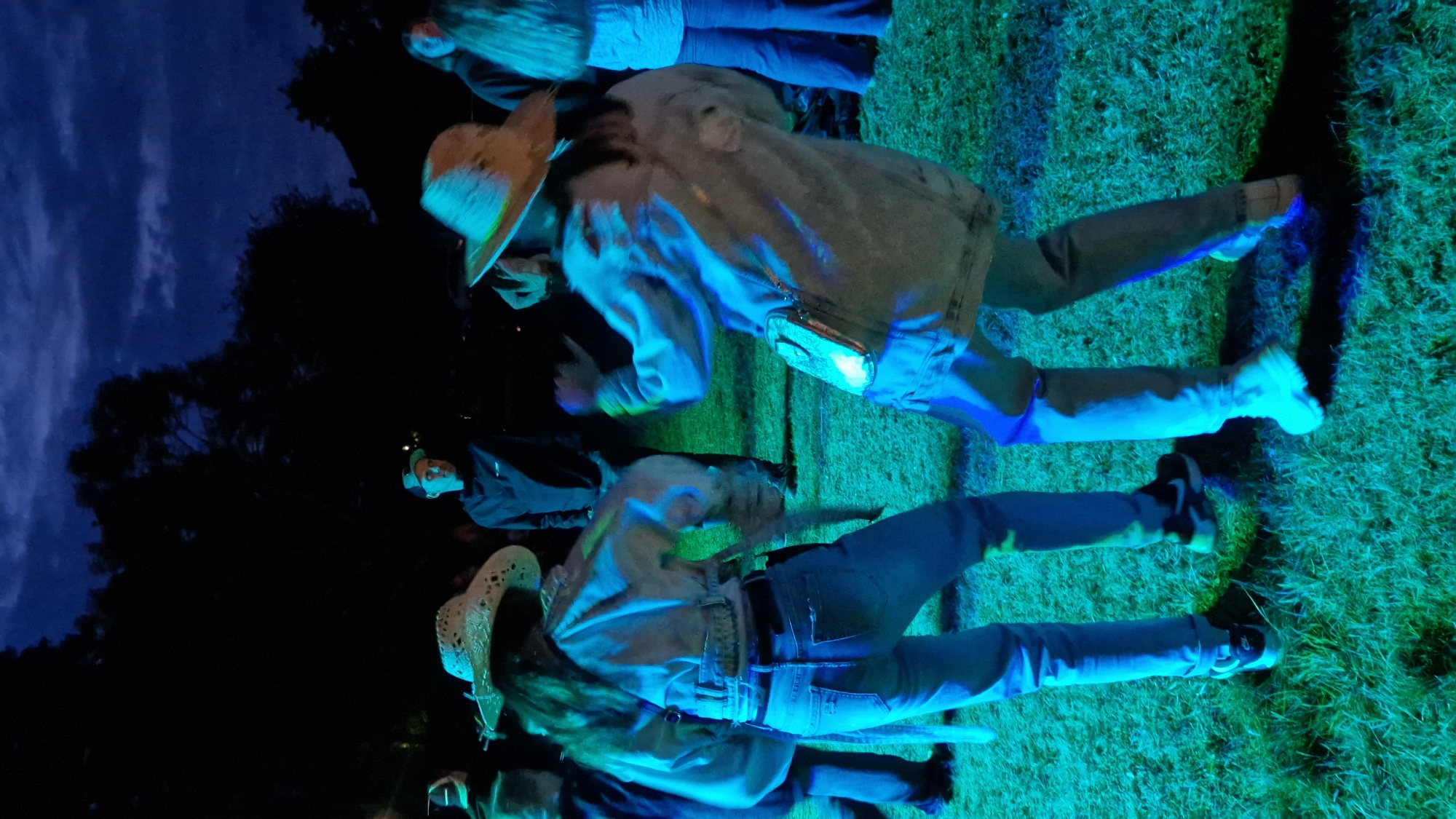 At dusk in Priekule, Latvia, two young women are dancing outdoors on a grassy area, illuminated by vibrant green and blue lights. Both are wearing denim jackets and jeans, and straw hats. The woman on the left has long brown hair, and the woman on the right has her back to the camera. In the background, other individuals are visible, standing and watching, or also dancing. Trees and a dark, cloudy sky suggest the event is taking place in a park or open field. The overall atmosphere appears festive and energetic.