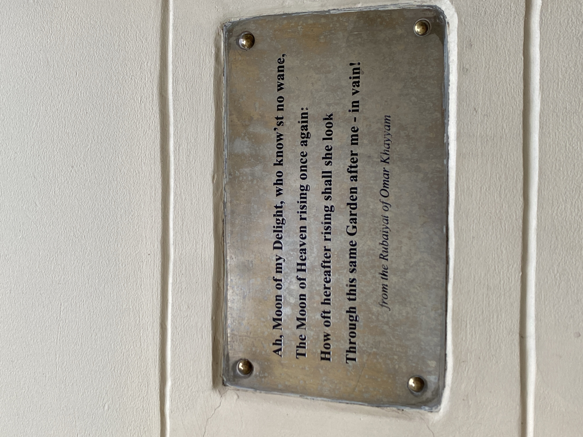 A close-up, eye-level shot shows a rectangular bronze plaque affixed to a textured, cream-colored wall in Kathmandu Metropolitan City, Nepal. The plaque is mounted with four visible brass screws, two on the top and two on the bottom edge, with the lower right screw reflecting light. The text on the plaque is in a dark, serif font. The inscription reads: "Ah, Moon of my Delight, who know'st no wane, / The Moon of Heaven rising once again: / How oft hereafter rising shall she look / Through this same Garden after me - in vain! / from the Rubaiyat of Omar Khayyam". The wall exhibits horizontal lines or indentations, suggesting it is constructed of brick or panels. The lighting is even, indicating an overcast day or shade.