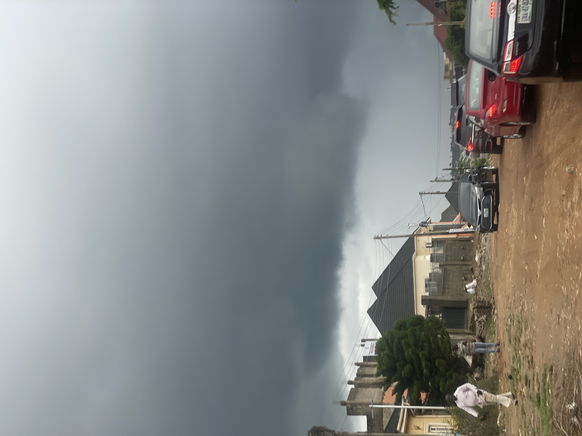 The image depicts a street scene in Abuja, Nigeria, under a dark, overcast sky indicative of impending rain. A dirt road runs through a residential area with houses of varying architectural styles lining the sides. Several cars are present, including a black sedan, a red sedan, and a dark SUV, suggesting traffic on the road. Two individuals are visible walking on the left side of the road, one in a light-colored shirt and trousers, and another in a striped shirt and jeans. The environment appears somewhat unkempt, with visible construction debris and vegetation. The overall atmosphere is somber due to the dramatic cloud cover.