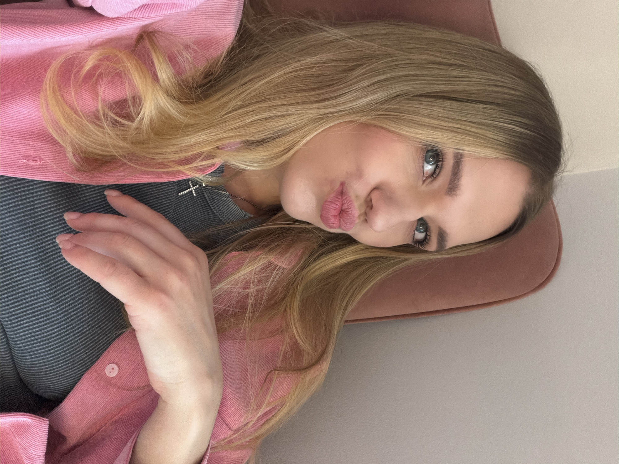 A young, fair-skinned woman with blonde hair is photographed from an elevated angle. She is wearing a pink corduroy jacket over a gray, horizontally-striped top, and a silver cross necklace. Her lips are pursed in a pouting expression. She is partially reclined on a rose-colored velvet surface. The background is a plain, light beige color. It is not possible to verify the location of Riga, Latvia with the visual information provided.