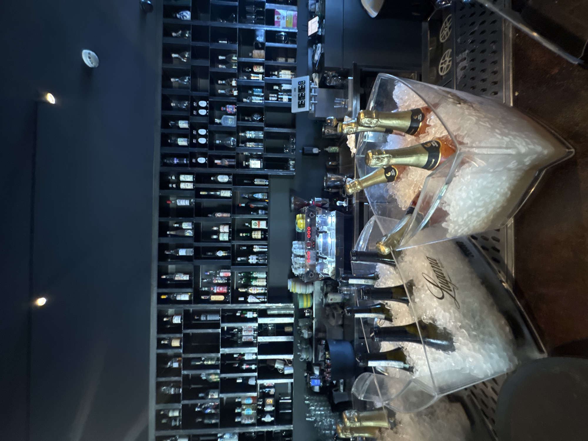 This media file depicts a well-stocked bar or restaurant interior, likely in Desenzano del Garda, Italy. The scene is dominated by a large, dark wall unit filled with rows of wine and liquor bottles, suggesting a sophisticated establishment. The bottles are neatly arranged in individual cubbies. In the foreground, a clear plastic ice bucket is filled with crushed ice and several bottles of champagne or sparkling wine, some with gold foil tops. The ice bucket has "Lugana" written in script across it. To the right, a metal bar top with a grated surface is visible, and behind it, a portion of a coffee machine with red lights is partially obscured.
The lighting suggests it might be evening or late afternoon, creating a moody ambiance. There are no people visible in the frame. Notable details include the ambient lighting which casts a soft glow, highlighting the bottles and the ice. The overall impression is one of a refined and possibly upscale venue, designed for serving beverages. The "Lugana" text on the ice bucket indicates a connection to the Lugana wine region, which is located near Lake Garda, further supporting the suggested Italian location.
