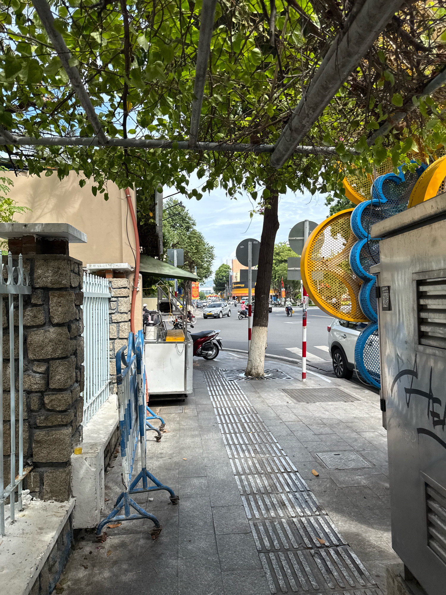 The image depicts a street scene in Nha Trang, Vietnam, on a cloudy day. In the foreground, a stone pillar with a metal fence stands to the left, partially obscured by an overhead vine-covered metal trellis. A blue and white metal barrier is positioned on the sidewalk. The sidewalk itself is paved with gray tiles and features tactile paving for the visually impaired. To the right, a large, metallic, cylindrical structure with yellow and blue decorative elements is visible, along with a white vehicle. The street beyond shows traffic, including cars and motorcycles, with buildings and trees lining the sides. A red and white striped pole is also present on the street. The overall impression is of an urban environment with ongoing activity.