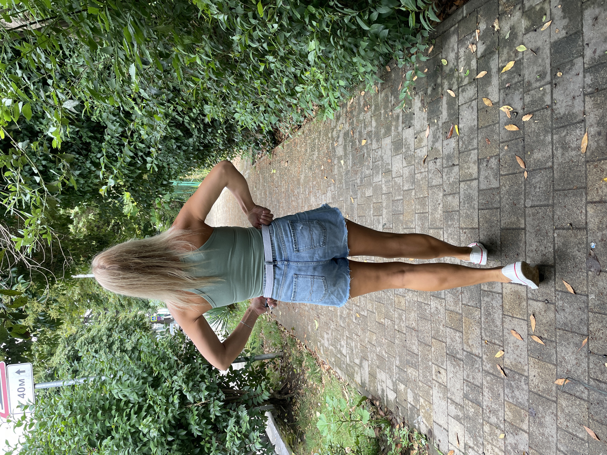 A Caucasian woman with long, blonde hair walks away from the camera on a paved pathway lined with green bushes and trees. She wears a light green tank top, denim shorts, a white belt, and white sneakers. A road sign indicating "40m" with an upward arrow is visible to her left. The pathway is made of rectangular paving stones, with fallen leaves scattered on it. The scene appears to be in an outdoor, possibly urban, park or street in Sochi, Russia.