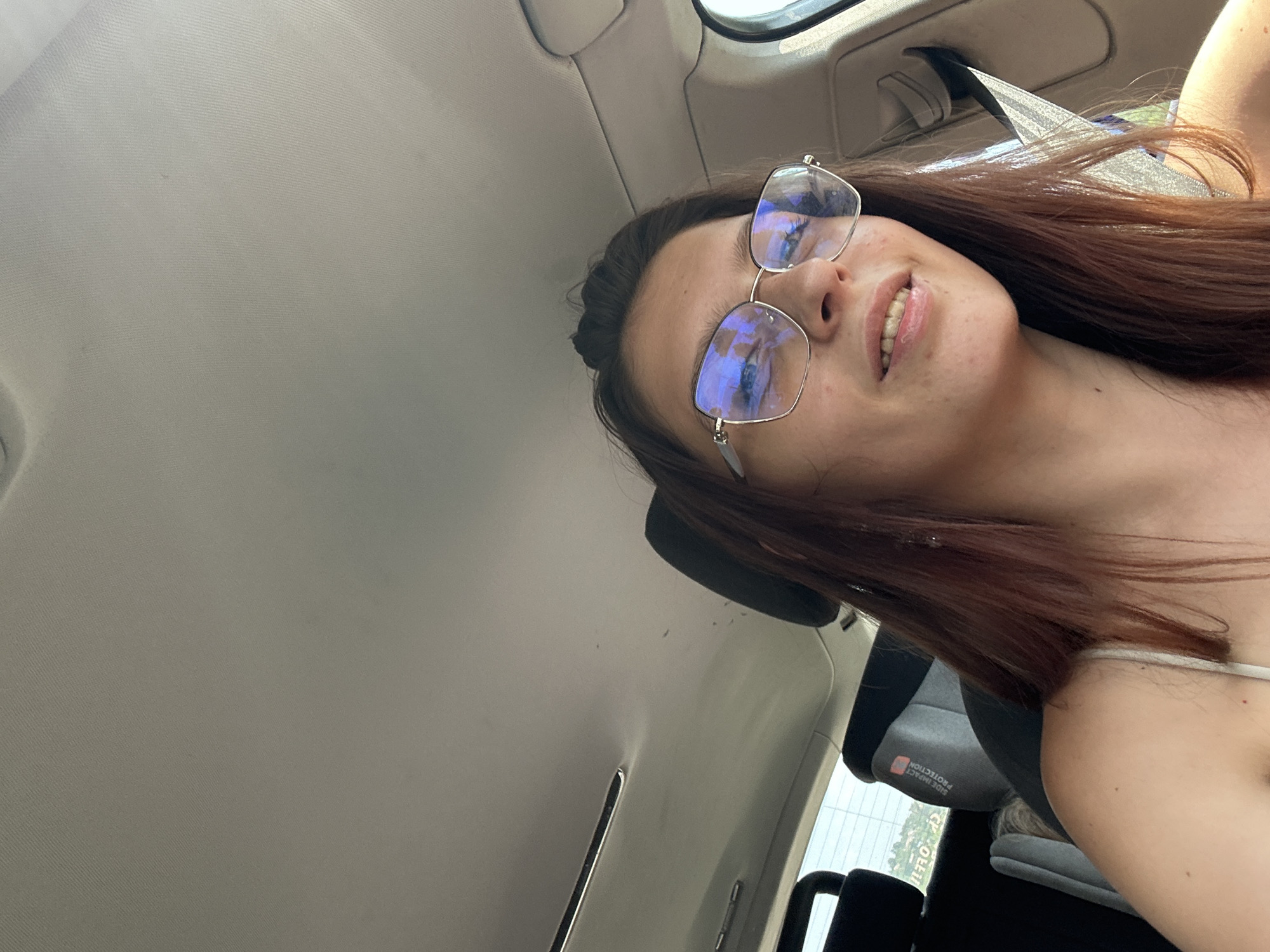 A young woman with long brown hair and glasses is sitting in the passenger seat of a car. She is wearing a light-colored tank top. The interior of the car is visible, including the ceiling and a headrest. Outside the car window, greenery and a road are visible. The image is a selfie taken from a low angle. The city of Tryavna, Bulgaria, is not depicted in this image.