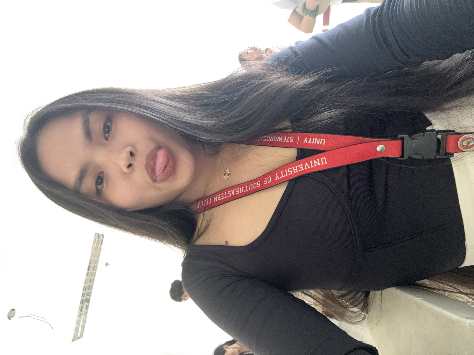 A young woman with long black hair is posing for a selfie, sticking her tongue out. She wears a black, long-sleeved shirt and a red lanyard with "UNIVERSITY OF SOUTHEASTERN PHILIPPINES" printed on it. The lanyard also has the words "UNITY | STEWARDSHIP". She has a clover-shaped necklace. The location is possibly Davao City, Philippines, due to the university name on the lanyard. Other people are faintly visible in the background. The environment appears to be indoors, possibly a school hallway.