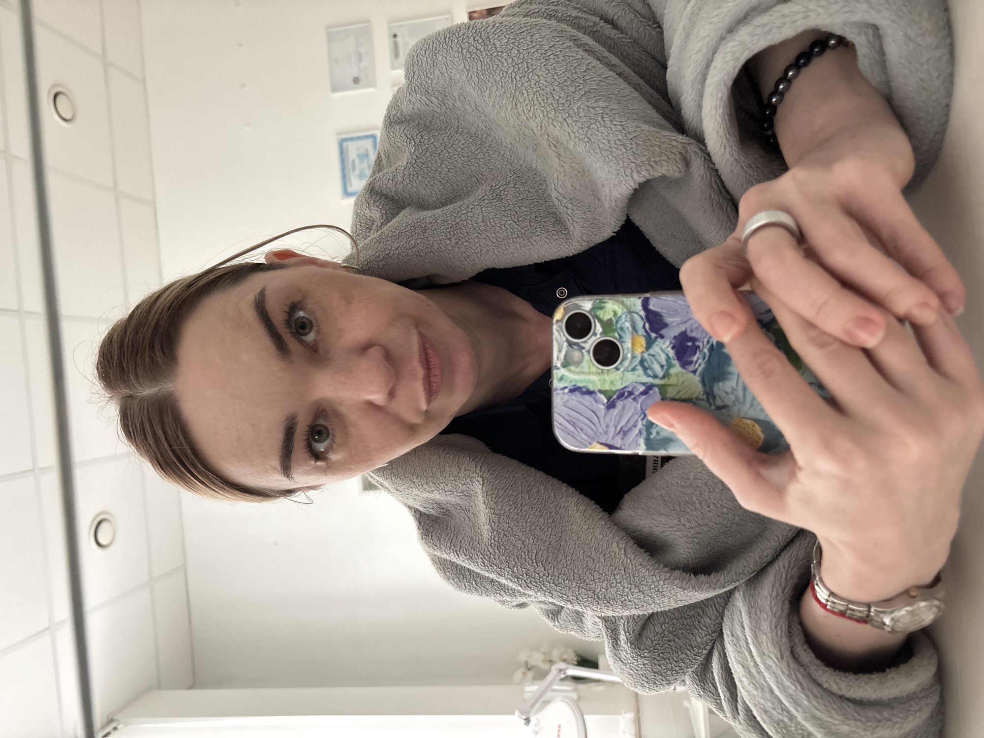A fair-skinned woman with light brown hair is holding a cell phone up to take a picture of herself. She is wearing a gray fleece robe and a dark blue top. She has a silver band on the ring finger of her left hand and a silver watch with a red string on her left wrist. A bracelet with black beads is visible on her right wrist. The background appears to be a tiled bathroom with a white tile wall and ceiling with recessed lighting. Certificates are visible on the wall behind her.