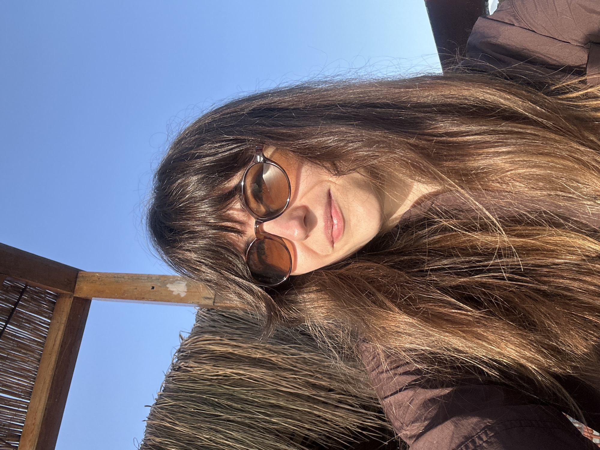 A woman wearing sunglasses poses for a selfie under a thatch-roofed structure. The sky is clear and blue. A wooden support beam is visible behind the thatch roof. The woman wears a brown shirt. Her hair is long and brown. The location is likely Alanya, Turkey, given the prompt's request to identify the location if possible.