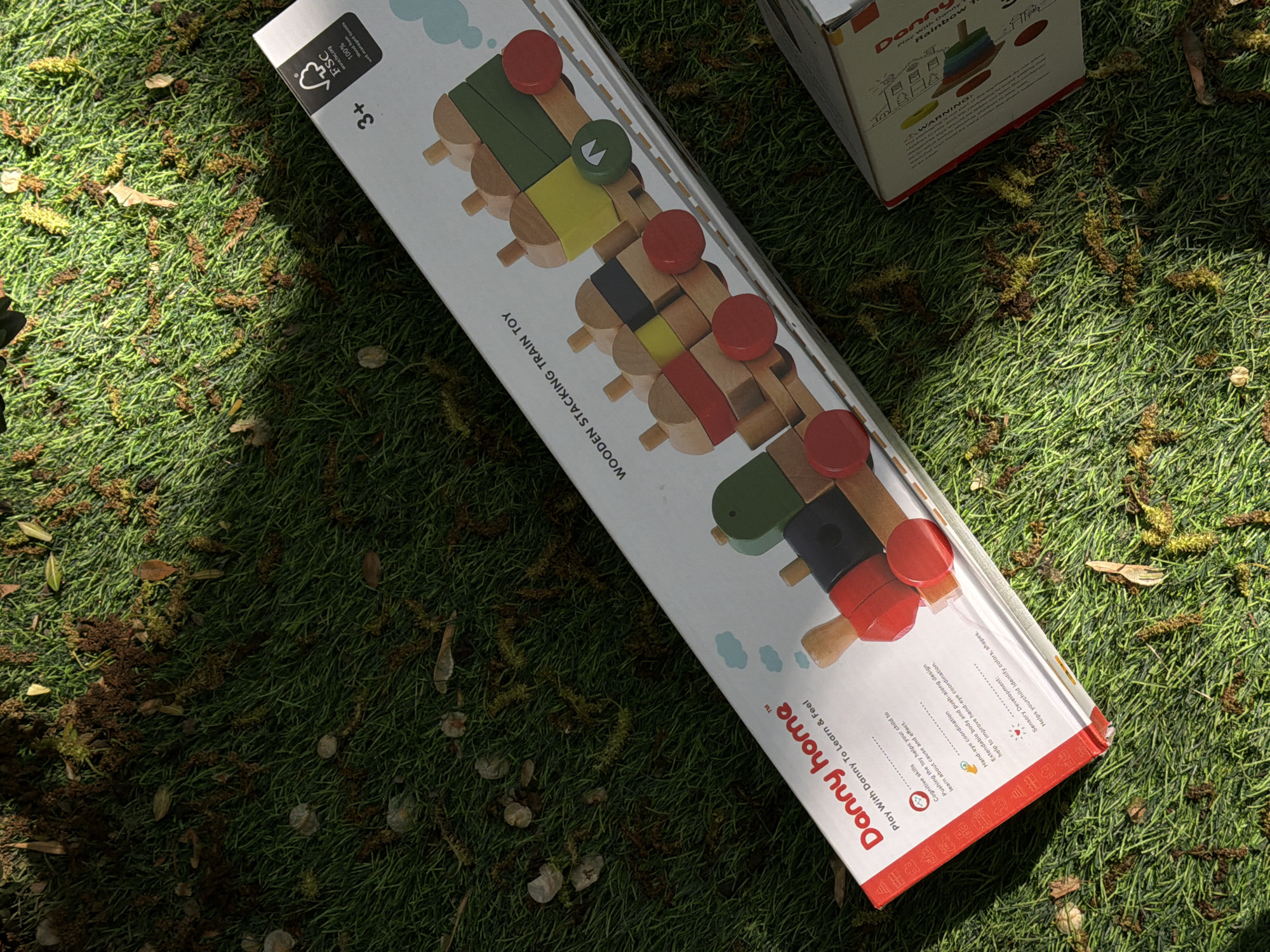 A box for a "Danny home" Wooden Stacking Train Toy sits on a surface that appears to be grassy. The box is white with red accents and features an image of a toy train constructed from colorful wooden blocks. The phrase "Play With Danny To Learn & Feel" is printed on the box, along with other details about the toy's educational benefits. A small part of another similar box is visible in the upper right corner. The location cannot be identified from the image.