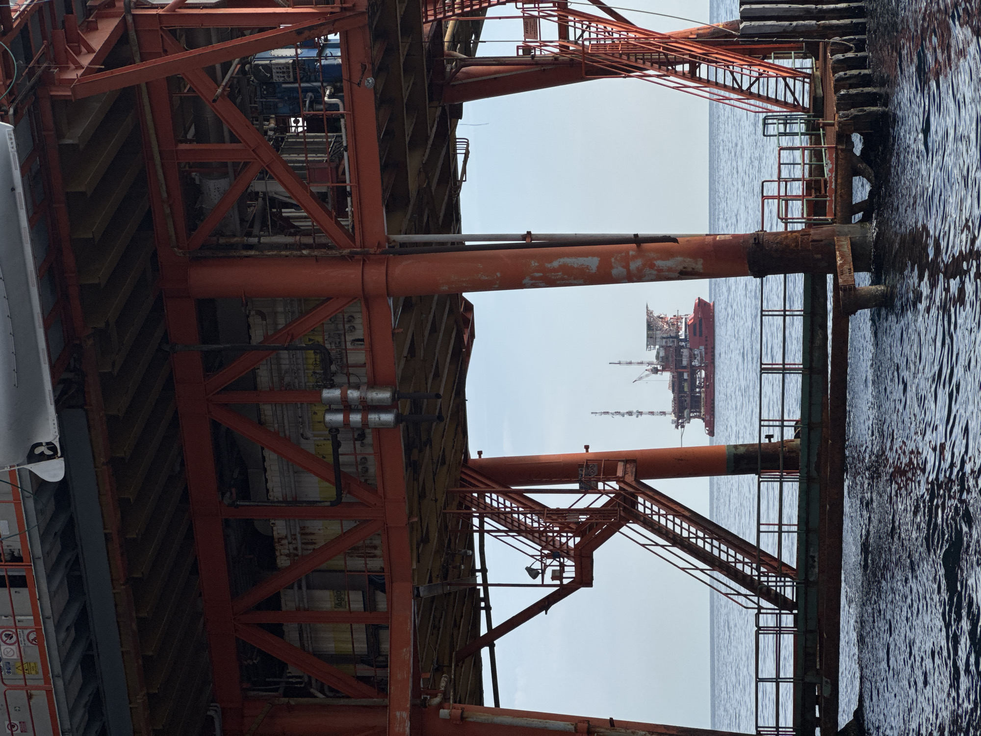 An offshore oil rig structure with red steel beams and platforms is in the foreground, with its support legs extending into the dark blue water. Metal staircases with railings are attached to the structure, leading up to various levels. In the background, a large red vessel, possibly another oil rig or a supply ship, is anchored on the calm, blue sea under a pale blue sky. The overall scene depicts an industrial maritime environment.