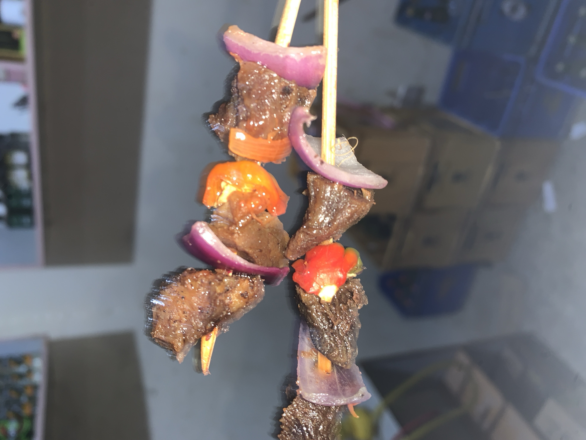The image shows two wooden skewers with pieces of cooked meat, red onions, and orange bell peppers. The skewers are arranged vertically, with the ingredients threaded in an alternating pattern. The background is blurry but appears to show an interior scene, possibly a home or restaurant, with items like boxes and containers visible. There is no indication in the image that it is located in Kabong, Nigeria, nor any details about people or interactions.