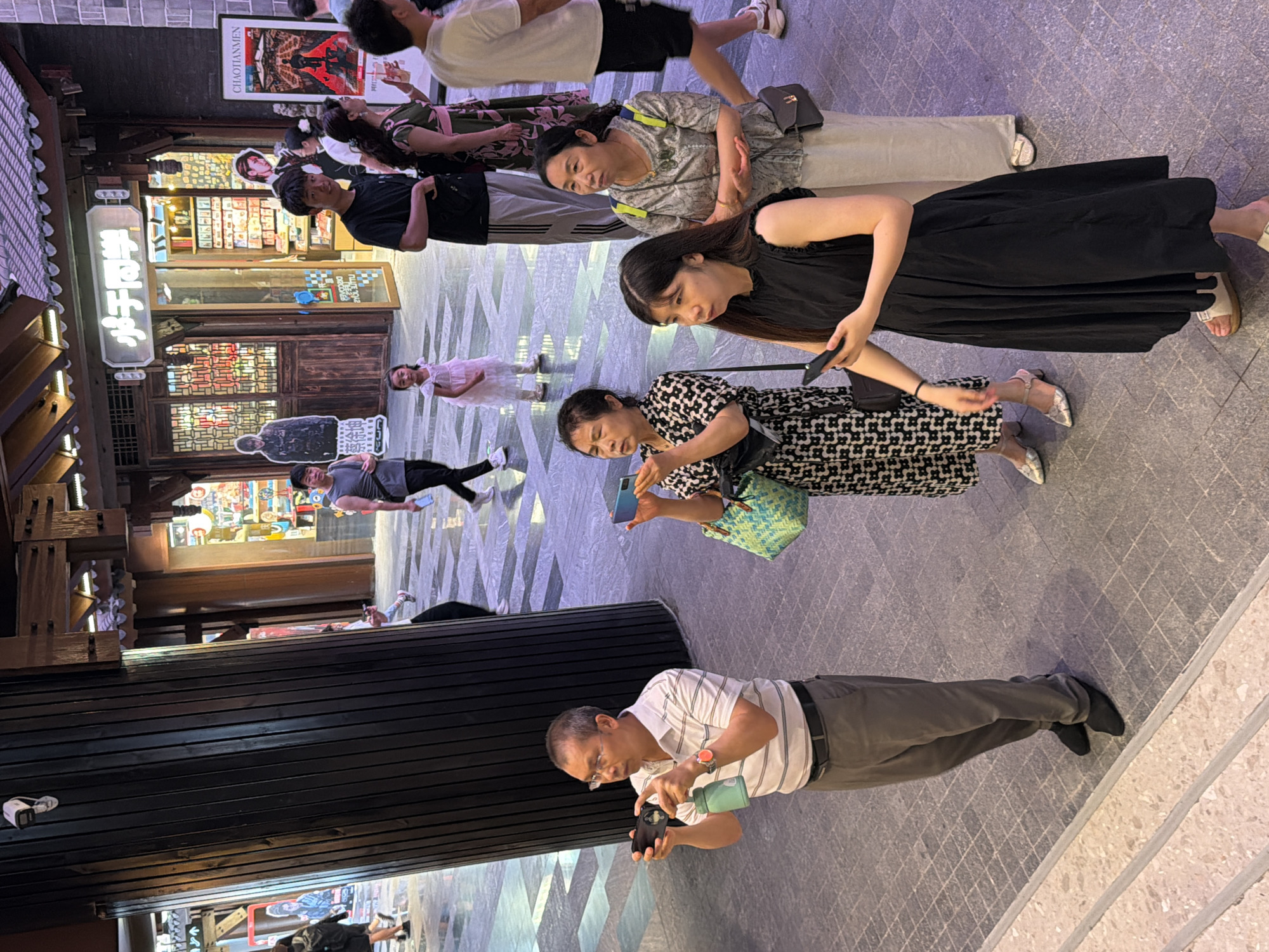 The image depicts a street scene in Yuzhong District, China, possibly a pedestrianized area or market. In the foreground, a man in a striped polo shirt and grey trousers holds two smartphones and a green water bottle. To his right, a woman in a black and white patterned dress holds a smartphone and a turquoise bag. Another woman, wearing a grey and white patterned top and a cream skirt, stands slightly behind and to the right, looking upwards. A young woman in a black sleeveless dress stands in the front right, holding a smartphone. In the background, several other people are visible, including a young girl in a pink dress. The architecture suggests a traditional Chinese style, with wooden beams and ornate detailing on the buildings. Shops with signs are visible, including one with Chinese characters and another advertising "CHAOTIANMEN". The ground is paved with large, light-colored tiles. The lighting suggests it is daytime, possibly with artificial lighting from the shops and overhead structures.