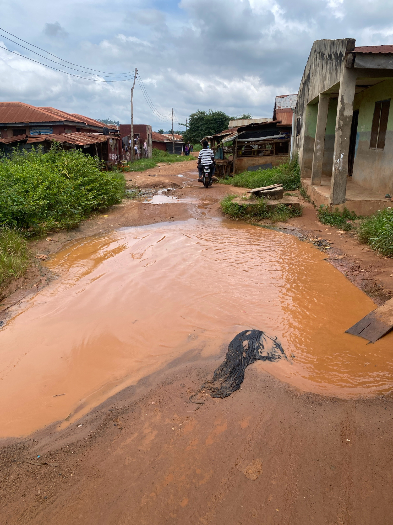 The image depicts a flooded dirt road in Iworoko, Nigeria, after rainfall. A person rides a motorcycle through a large, muddy puddle that covers most of the road. To the right, a weathered building with a green facade stands adjacent to the road. On the left side, several structures with corrugated metal roofs are visible, along with dense green foliage. The sky is overcast with clouds. A dark object, possibly debris, is partially submerged in the muddy water.
