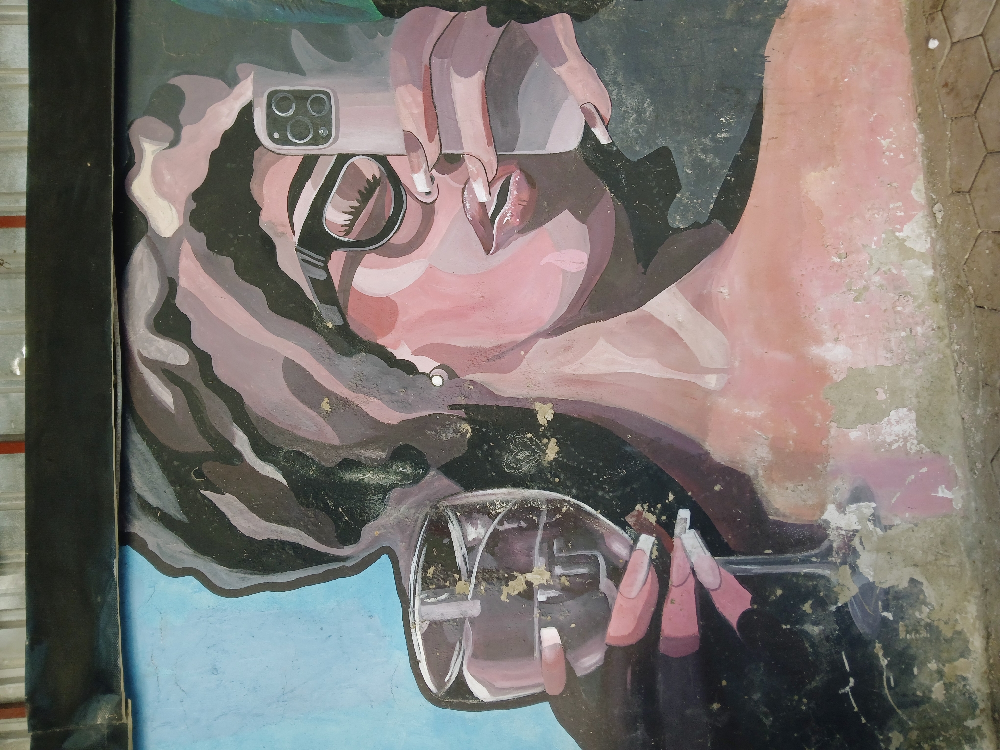 The image shows a mural depicting a stylized portrait of a person holding a cell phone and a glass. The mural appears to be painted on a weathered wall in a distressed condition, with visible peeling paint and surface damage. The portrait is rendered in shades of pink, grey, and black, with simplified shapes and lines. The person is holding a cell phone with multiple camera lenses up to their eye as if taking a picture. They are also holding a glass, possibly a wine glass. The background is a combination of blue and dark grey areas. The exact location in Barakin Lerawi, Nigeria is not discernible from the image.