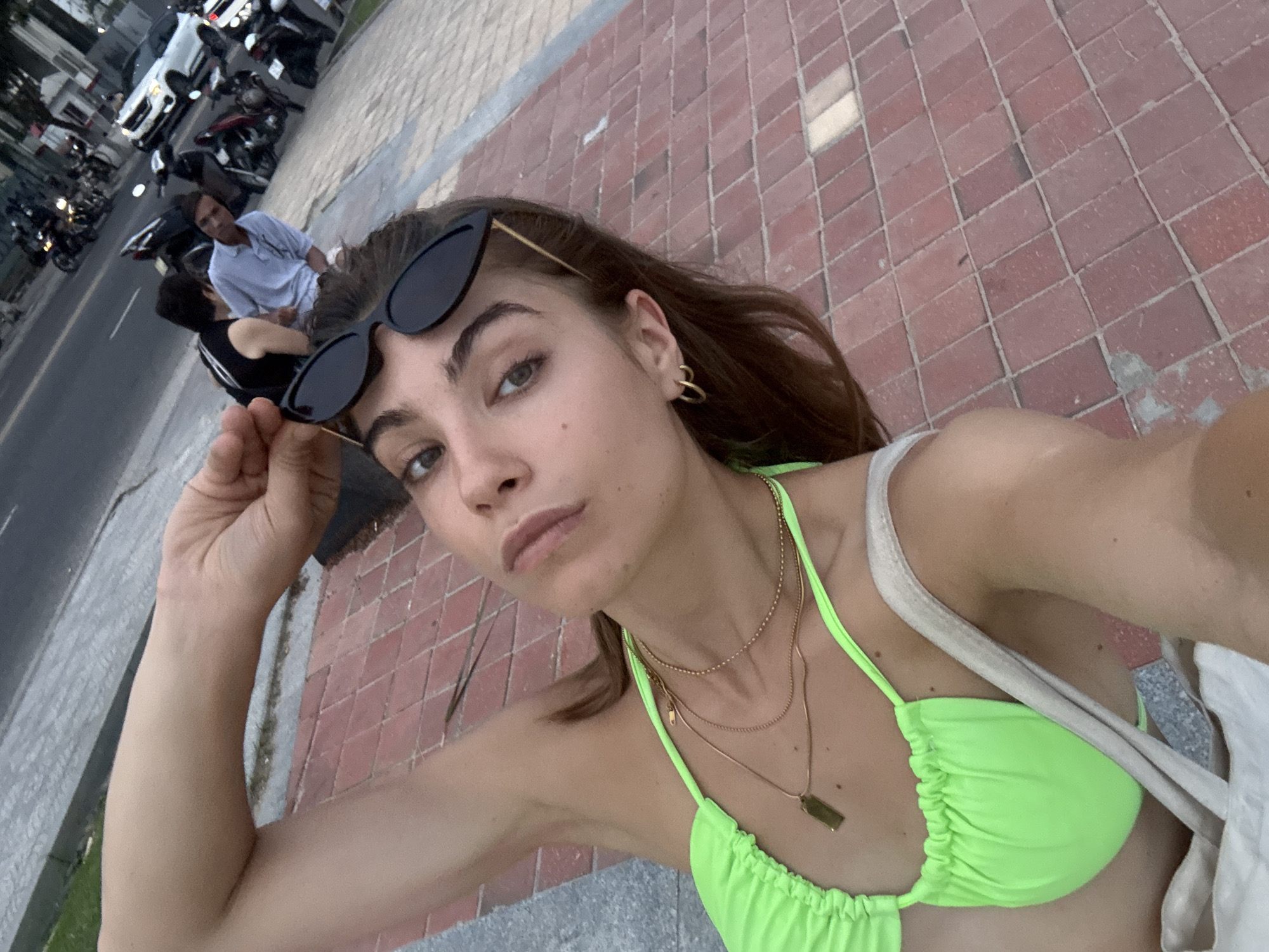 A woman is taking a selfie while standing near a brick-paved area. She is wearing a neon green bikini top and has sunglasses resting on her head. She is fair-skinned, with dark hair, and wearing multiple gold necklaces. In the background, there are people sitting on scooters parked along the street, and cars and scooters are also visible. The background setting indicates the location is in the city of Đà Nẵng, Vietnam.