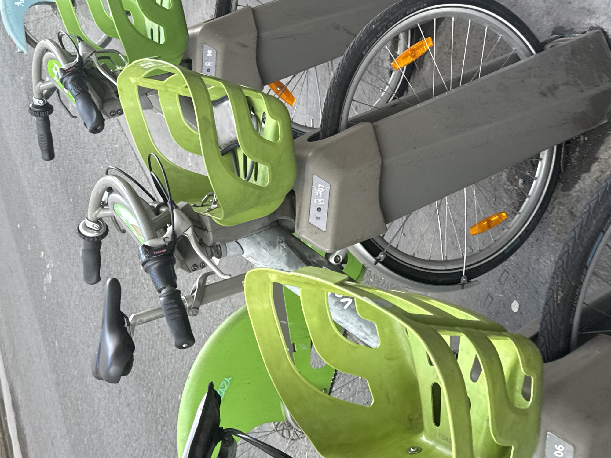 The image displays a close-up, rotated view of several lime-green public rental bicycles docked in a grey metallic station, characteristic of Vélib' Métropole in Paris, France. The bikes are neatly aligned in their respective docking slots, suggesting they are available for rent or have recently been returned.
The primary subjects are the bicycles and their docking infrastructure. Each bicycle features a distinctive lime-green frame, front basket, mudguards, handlebars with black grips, and seats. Visible details on the handlebars include a "Nexus" brand shifter, indicating internal gear hubs. The wheels have silver spokes, black tires, and bright orange reflectors.
The docking station is constructed from grey metal, with individual slots for each bike. On the side of the docking stations, numbers like "0-07", "06", and a partially visible "08" are present, likely identifying the specific dock numbers. The overall scene is outdoors, likely on a paved street or sidewalk, under clear daylight conditions, given the consistent natural lighting and lack of shadows indicating late evening or night. No people are visible in the frame, focusing entirely on the parked bikes.
Faint, possibly graffiti-like markings are visible on the green mudguards of some bicycles, and the word "ENIAS" is partially discernible on one of the green front baskets. The image is taken from a slightly elevated, overhead perspective, with the entire composition rotated 90 degrees clockwise.