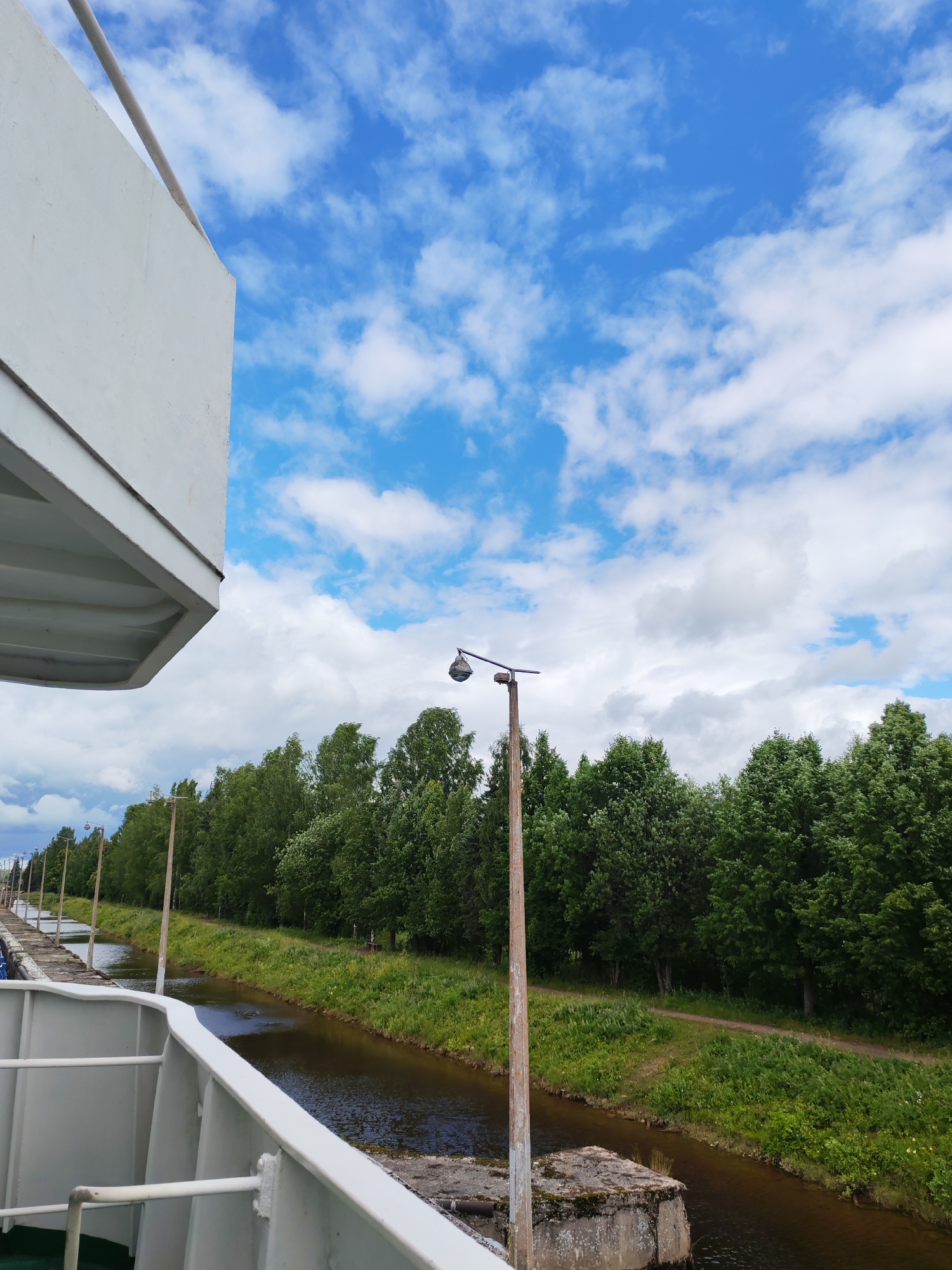 The image, taken from a vessel on the Vytegra River in Vytegra, Russia, shows a canal with lush green trees lining its banks. The sky is a vibrant blue with scattered white clouds. In the foreground, a white railing of the vessel is visible, along with part of its structure. A wooden pole with a light fixture stands near the canal's edge, and a dirt path runs parallel to the water. The scene suggests a peaceful day for navigation on the river.