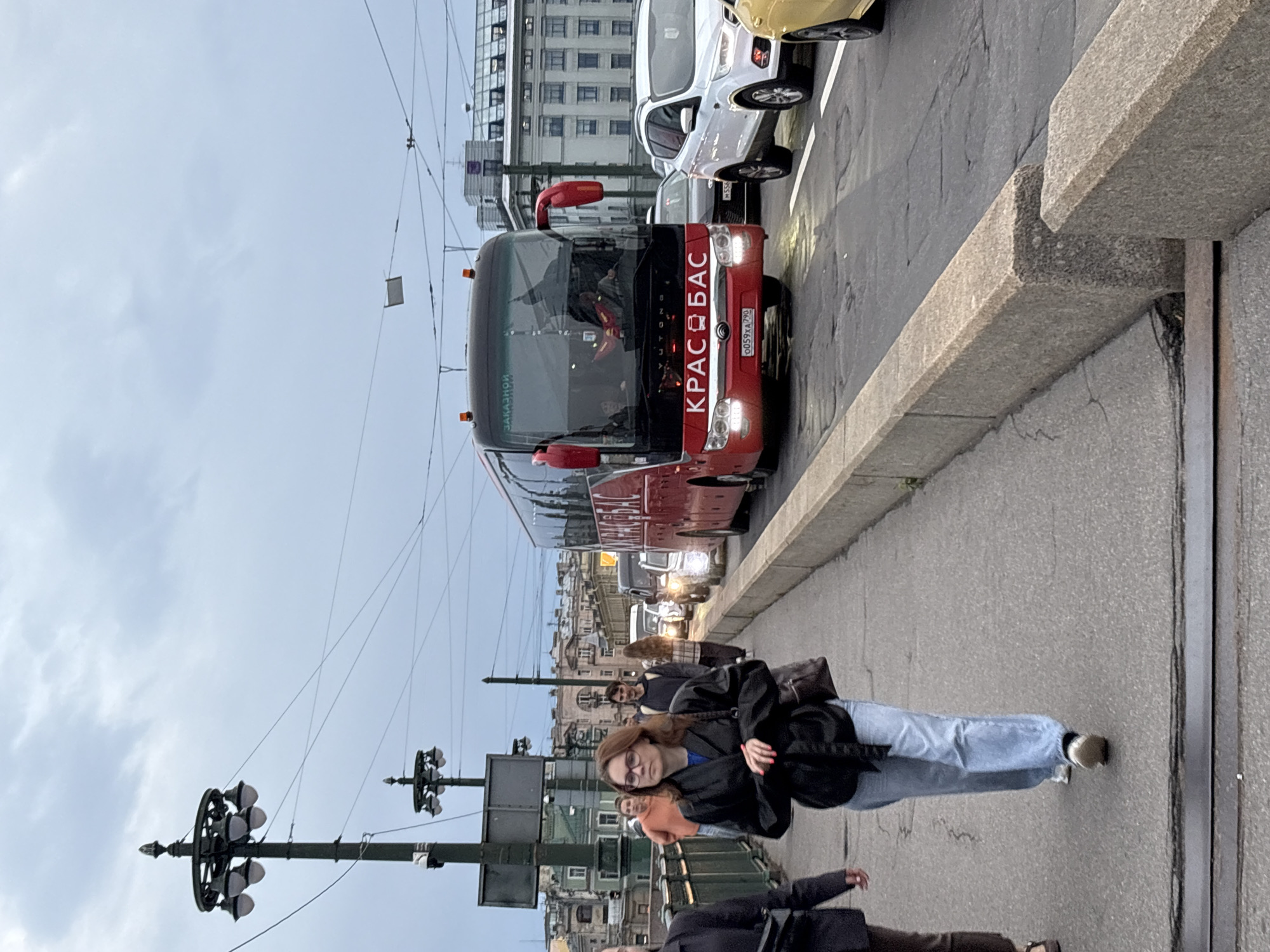 A person with glasses and brown hair is walking on a sidewalk next to a busy street. They are wearing a black jacket, a blue shirt, and light blue jeans. The person is looking at the camera. In the background, there is a red bus with "КРАСАВАС" written on the front in white letters. The bus has a license plate that reads "0059XA 790". There are other cars on the street, and buildings in the distance. The sky is overcast, suggesting it might be a cloudy day. The scene appears to be in Saint Petersburg, Russia, given the architectural style of the buildings and the style of the bus. The overall mood is casual, with people going about their day. There are streetlights and electrical wires visible, indicating an urban setting.