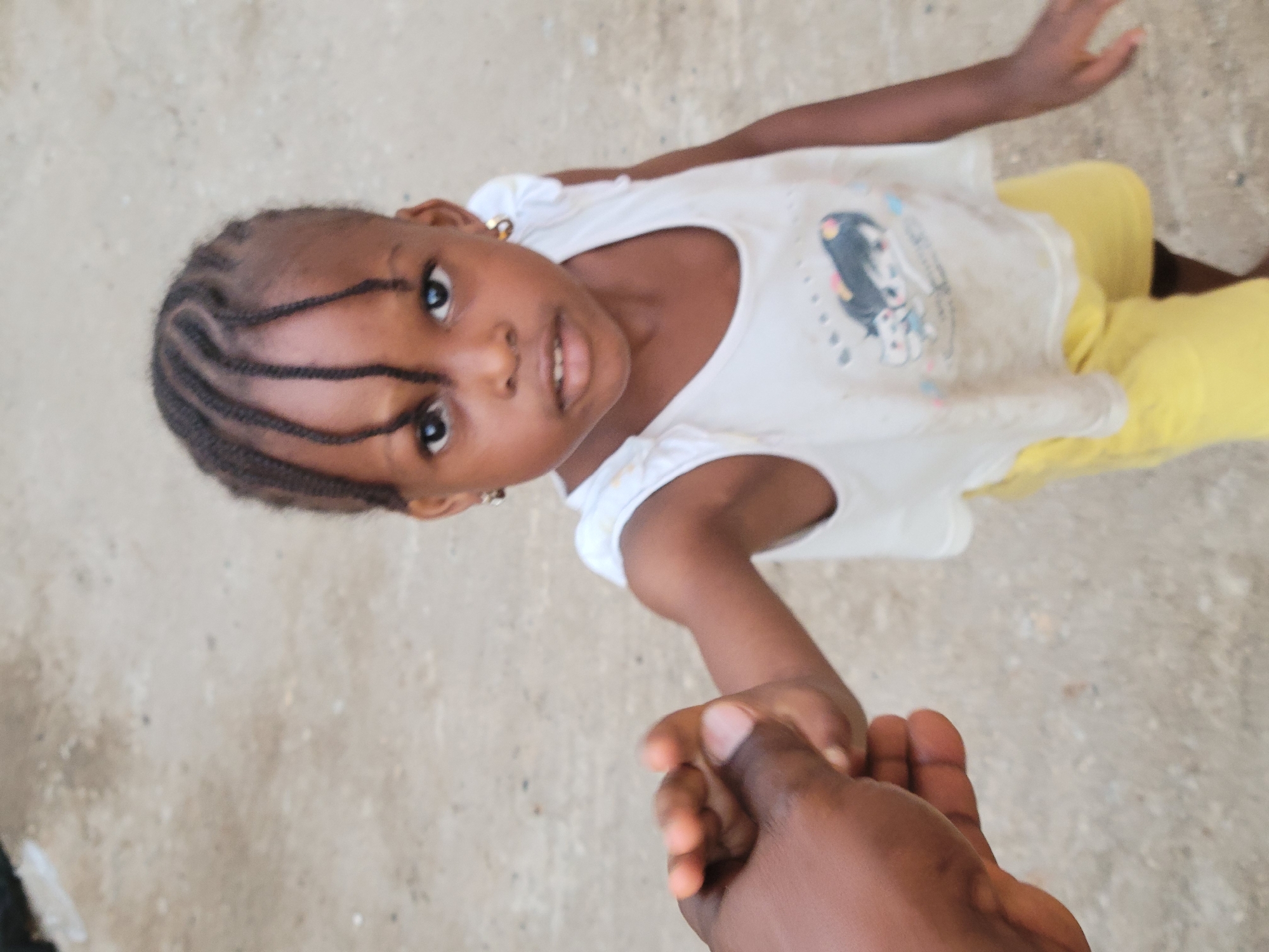 A high-angle, close-up photograph captures a young Black girl holding hands with an unseen adult, looking upwards towards the camera. The image is rotated, with the girl appearing diagonally from the top left towards the bottom right.
The girl, who appears to be a toddler or young child, has dark skin, dark eyes, and dark hair styled in cornrows or braids that sweep across her forehead. She wears small gold hoop earrings. Her expression is one of gentle curiosity or a soft smile, with her mouth slightly open. She is dressed in a white t-shirt featuring a light-colored graphic design of what appears to be cartoon characters, and bright yellow pants or shorts.
The hand of an adult, also with dark skin, is visible in the foreground at the bottom right, firmly holding the girl's hand.
The setting appears to be outdoors on a light brown, sandy, or dirt ground, suggesting an informal environment. The natural lighting indicates it is daytime. The overall scene conveys a moment of connection and tenderness between the child and the adult. No discernible text is visible on the girl's clothing beyond the general cartoon graphic. The location context provided is Madalla, Nigeria.