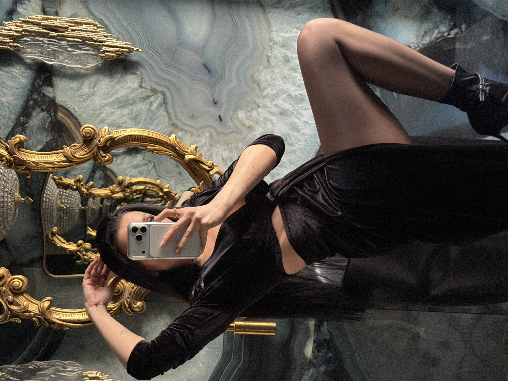A female subject is depicted taking a mirror selfie in a lavish interior, located within the city of Moscow, Russia. She wears a dark velvet two-piece outfit: a long-sleeved crop top revealing her midriff, and a skirt with a high slit. Her right leg, clad in dark hosiery and a black ankle boot with a silver buckle, is bent and elevated. Her right hand holds a silver smartphone, obscuring her face, while her left hand is raised near her head. Long, dark hair is visible. The background features a wall with a prominent blue, white, and grey veined stone or marble pattern. Two ornate, gilded mirrors with baroque-style frames are mounted on this wall, reflecting ceiling light fixtures. A gold-colored cylindrical fixture is visible on the wall to the subject's left, and a gilded, textured wall decoration is present to her right. A ring is visible on her right hand's ring finger, and her fingernails are painted a light shade.