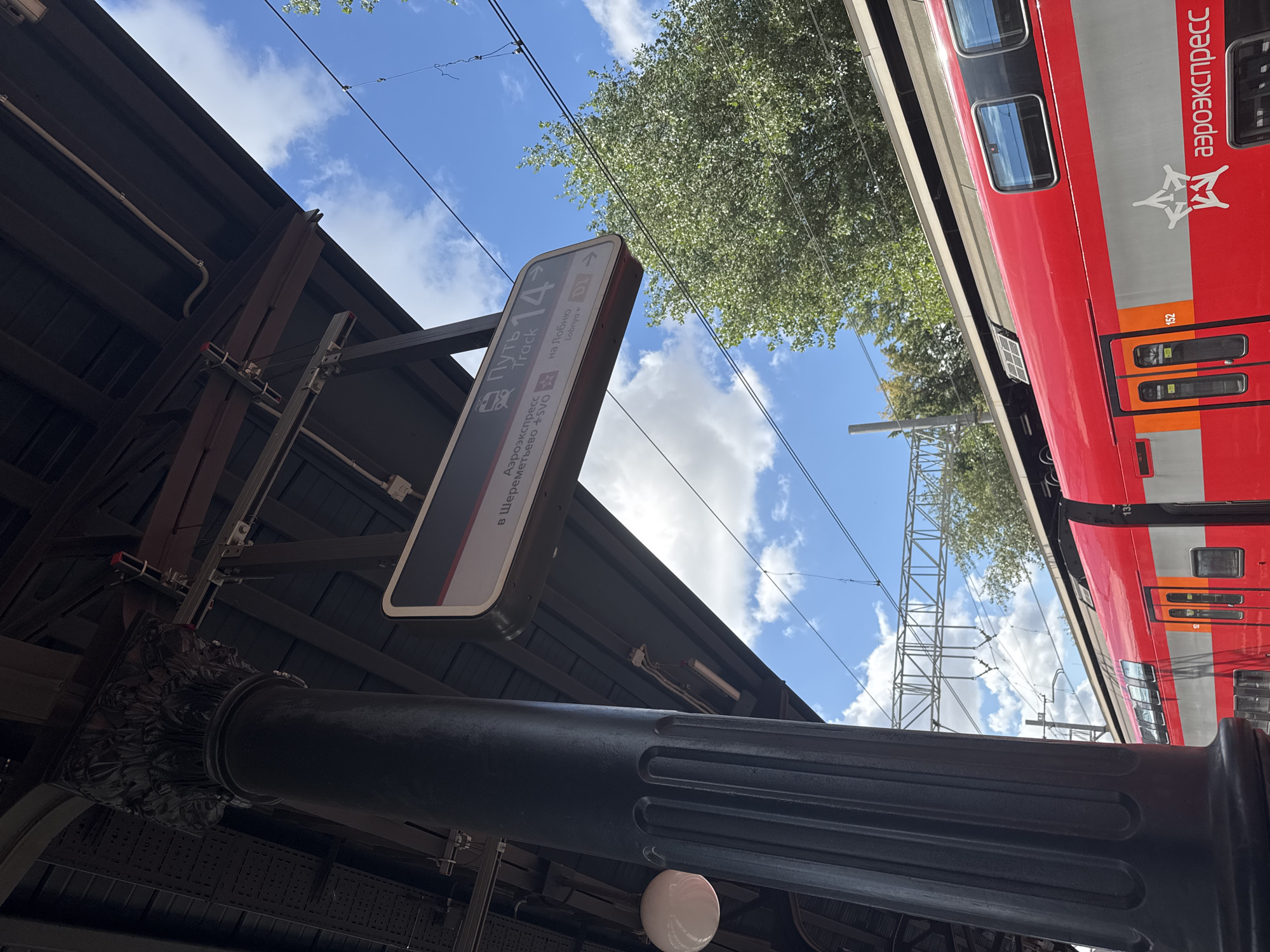 The image shows a close-up of a platform at a train station in Moscow, Russia, with a red and silver double-decker train pulled up to the platform. In the foreground, a large, dark, fluted column is visible on the left, part of the station's architecture. Above the column, a digital sign displays train information in Russian and English. The sign indicates "Aeroexpress," "Path Track 14," and lists destinations including "Sheremetyevo SVO" and "Lobnya." The train itself has multiple windows and doors, and the "Aeroexpress" logo is visible on its side. In the background, green trees and a bright blue sky with scattered clouds suggest a clear day. Overhead, electrical wires and support structures for the train tracks are visible. The overall scene conveys the operational atmosphere of a busy Moscow transport hub.
