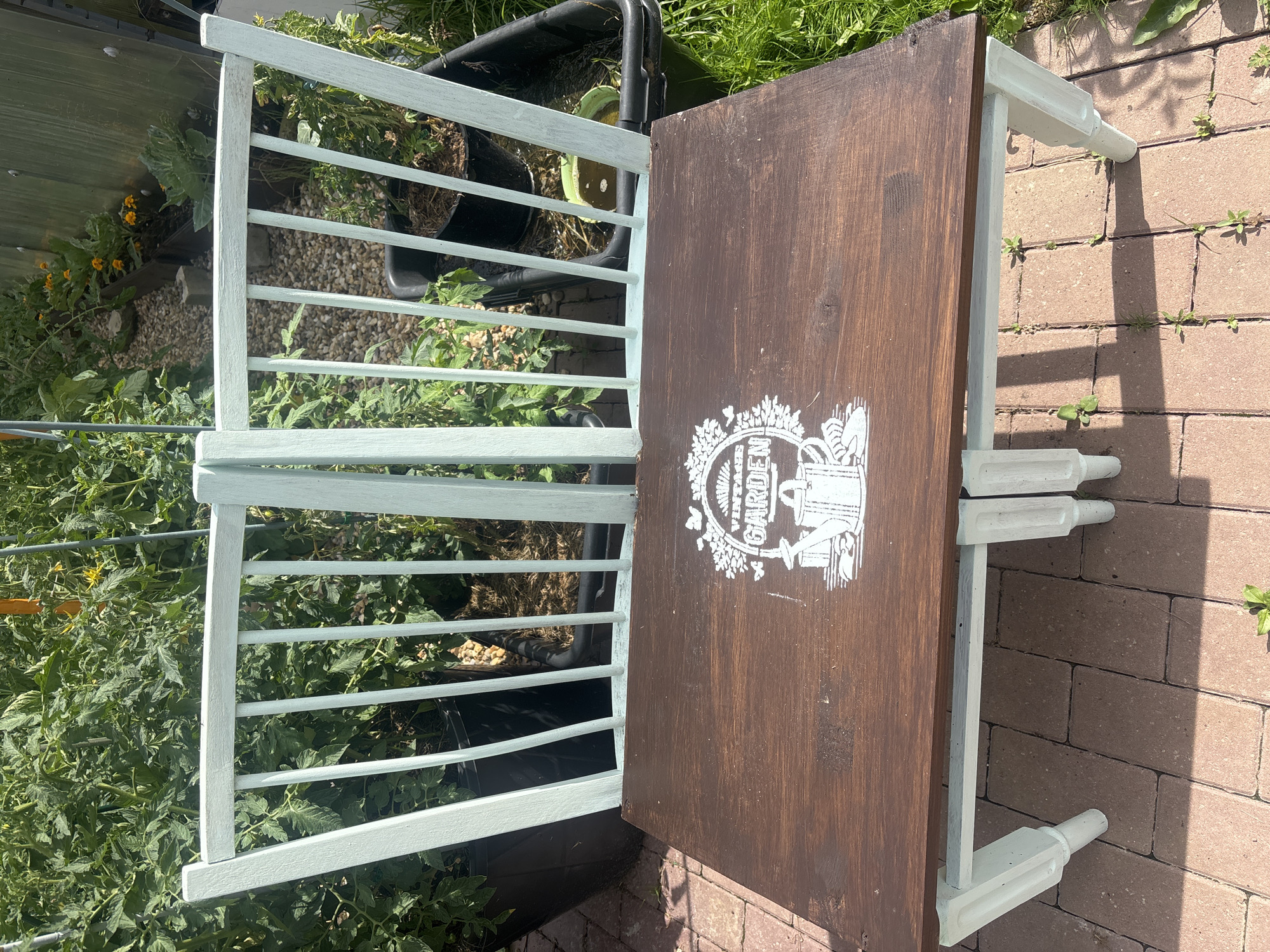 The image shows a wooden bench with a light blue painted frame and a dark brown seat. The seat is decorated with a white stencil that depicts a watering can, some plants, and butterflies, with the word "GARDEN" in the center. The bench is placed outdoors on a brick patio, with a garden in the background. The garden appears to be a vegetable garden with tall green plants, some of which are supported by stakes. There are also some black plastic planters and a wheelbarrow visible in the background. The lighting suggests it is a sunny day. The location is described as Lotosh SNT (Maryino), Russia.