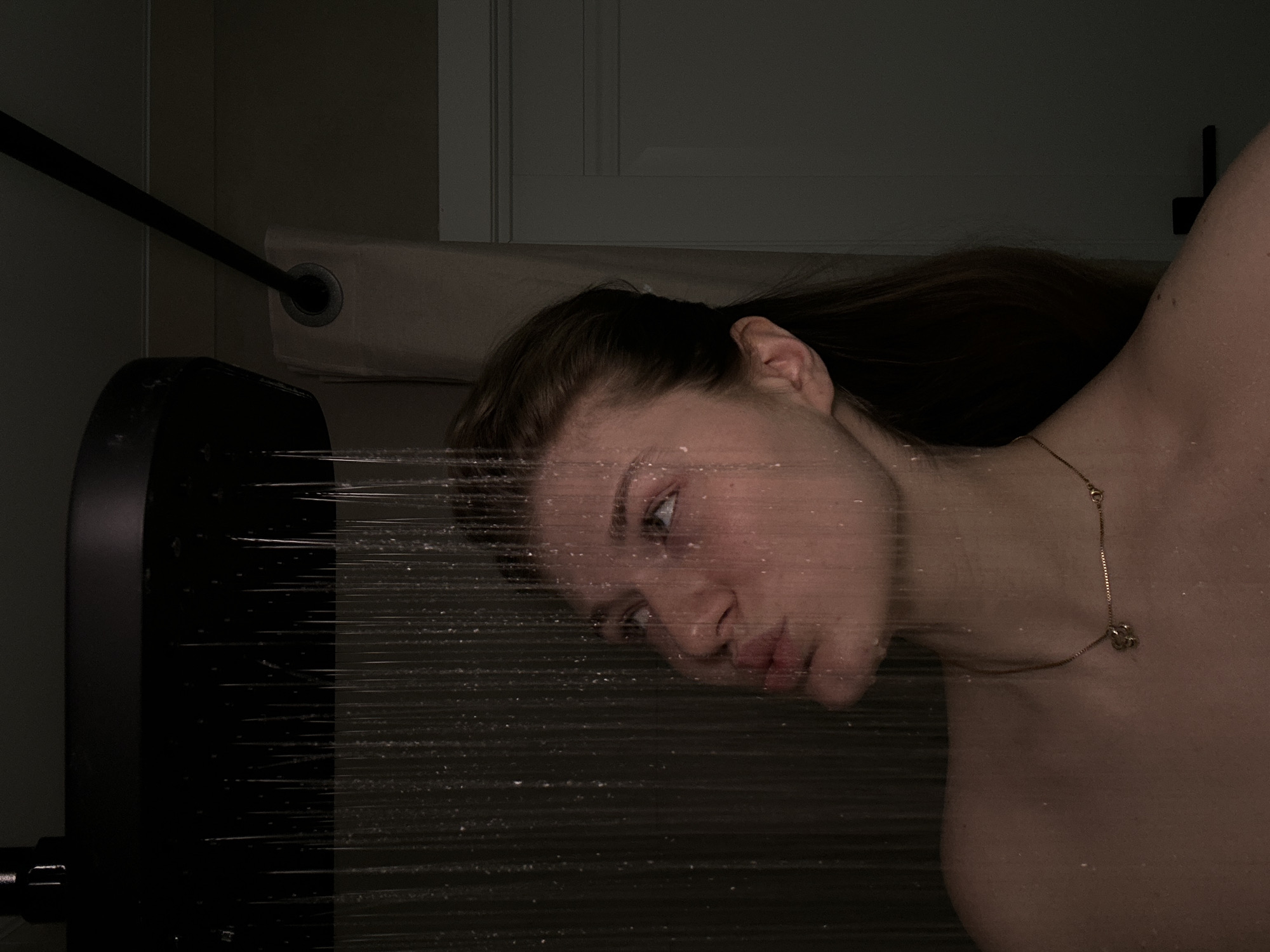 A portrait of a fair-skinned woman with light brown hair and a gold necklace. Her face is visible through clear plastic wrap. She is looking to the left of the frame. To the left of the woman's face is a dark object with horizontal lines running through it. In the background are a dark wall with a partially visible door and curtain rod. It is not possible to tell whether the location is in the city of Kazan, Russia.
