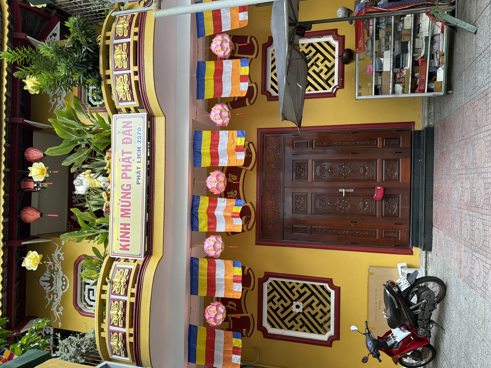 The image depicts the exterior of a vibrant yellow building, likely a temple or shrine, in Nha Trang, Vietnam. The facade features ornate architectural details, including a yellow and red tiled roof, decorative balconies with golden floral motifs, and intricate carvings around the entrance. A prominent sign above the entrance reads "KINH MUNG PHAT DAN PHAT LICH 2570" in pink lettering.
Several colorful Buddhist flags, in stripes of blue, yellow, red, white, and orange, hang across the front of the building, interspersed with pink lotus-shaped lanterns. Two large, dark brown wooden doors form the main entrance, flanked by square decorative windows with geometric black and white patterns. To the left of the entrance, a red and black motorbike is parked on a patterned tiled pavement. To the right, a glass display cabinet filled with various items, possibly merchandise or offerings, stands on the pavement. A portion of a dark umbrella is visible to the right of the cabinet. A woman with grey hair and wearing a patterned blue and white outfit is partially visible standing behind the display cabinet, appearing to interact with its contents. Red paper lanterns, one yellow and two red, are suspended from the upper part of the building, contributing to the festive and spiritual ambiance. Lush green plants add a touch of nature to the scene, growing from the balconies and near the entrance.