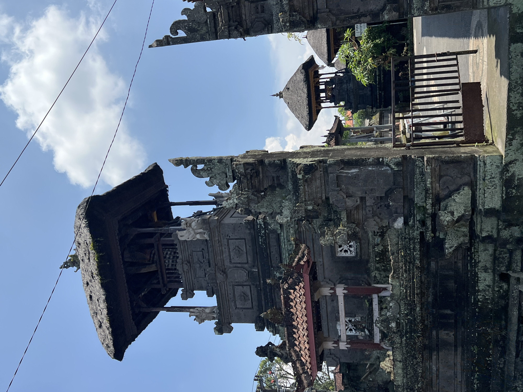 The image captures a detailed view of a multi-tiered Balinese Hindu temple structure in Sembung, Indonesia, under a bright blue sky with scattered white clouds. The dominant structure in the foreground is dark gray stone, extensively covered in green moss and lichen. Its uppermost section features a dark, weathered thatched roof with a small, carved finial. Below the roof, an open, square wooden pavilion contains a dark, vertical element, possibly a shrine or statue, flanked by white, stylized stone guardian figures at the corners. Lower tiers of the stone edifice display carved relief panels. To the immediate left, a smaller, lower structure with a gabled roof of reddish-brown tiles is visible, featuring white decorative elements and red and white pillars.
To the right of the primary structure, a traditional Balinese stone gateway, also heavily covered in moss, frames the view into the temple grounds. Through this gateway, a partially open, dark metal bar gate with rust on its lower section leads to an asphalt road or path. Beyond the gate, another smaller temple pavilion with a thatched roof is visible in the background, alongside green foliage and a white sign partially bearing the text "PURA PUSUH LAN." A single dark cable extends diagonally across the upper portion of the blue sky. No individuals are present in the depicted scene.