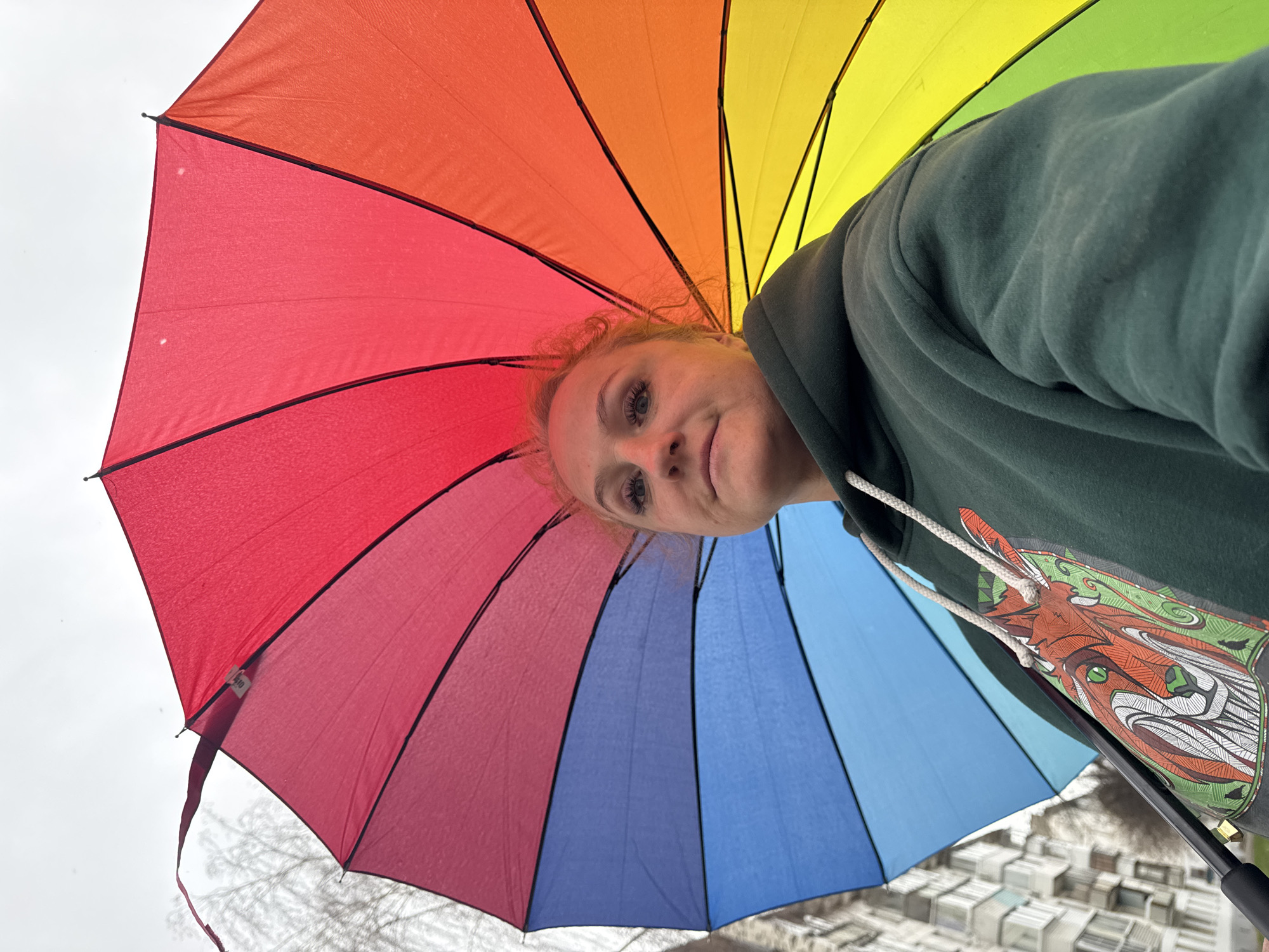 The image depicts a woman holding a rainbow-colored umbrella, looking down at the camera. She is wearing a dark green hoodie with a stylized animal design on the chest. The background shows a cloudy sky and a blurry cityscape. The location is Chaikovsky, Russia.