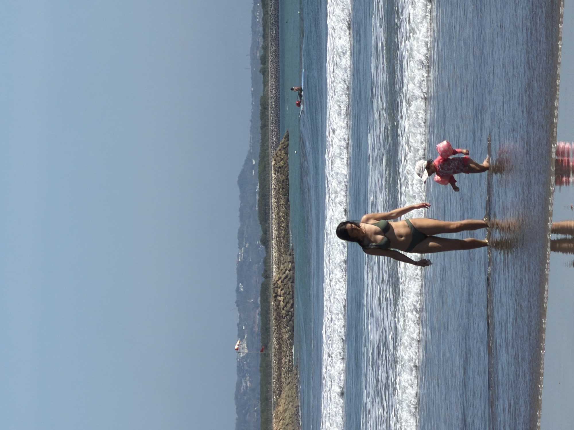 The image depicts a bright, sunny day at Kuta Beach in Indonesia. In the foreground, a woman in a green bikini stands in shallow water, with gentle waves lapping around her legs. Her body is positioned towards the camera, and she appears to be looking down and slightly to her left.
In the background, another person, possibly a child, is visible further out in the water, wearing a pink floatation device and a head covering. This individual is lying on their back in the water, with their arms outstretched.
The scene includes a long breakwater made of dark rocks extending into the ocean, separating the calm waters of the lagoon from the choppier sea beyond. In the distance, a hazy coastline with buildings and vegetation can be seen. The sky is clear and a bright blue, suggesting fair weather. The reflection of the water and the sky are visible on the wet sand, adding to the serene beach atmosphere. There is no visible text in the image. The overall mood is one of leisure and enjoyment of a tropical beach setting.