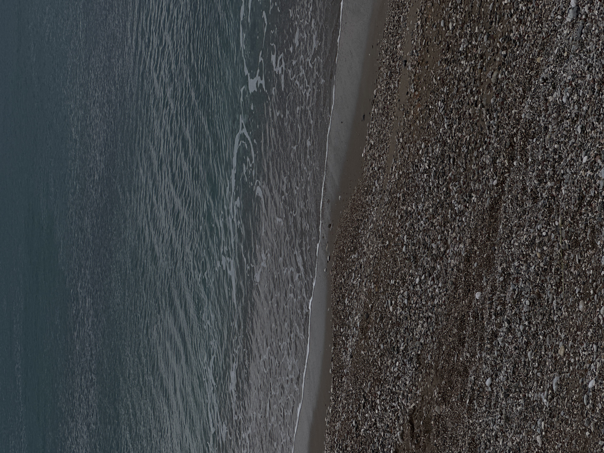 The image depicts a section of coastline, likely in Šušanj, Montenegro, characterized by the interface of the Adriatic Sea and a pebble beach. The left portion of the image shows the sea with subtle wave action visible. The water transitions from a deeper tone on the left to lighter tones near the shoreline, suggesting shallowing depth. A narrow strip of wet sand separates the water from a wider stretch of pebble-covered beach on the right. The pebbles range in size and color, forming a densely packed surface. The composition is a high-angle or aerial view. No people or specific objects are distinctly visible.