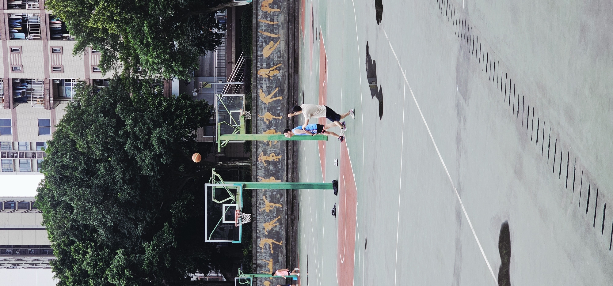 The image shows a basketball court in Yuzhong District, China, with two men standing near the foul line. One man is wearing a tan shirt and dark shorts, while the other is wearing a blue shirt and dark shorts. Both are facing towards the basketball hoop. A basketball is suspended in the air between the men and the hoop. A wall with figures painted on it is behind the men. There are puddles on the court, and a building with trees is in the background.