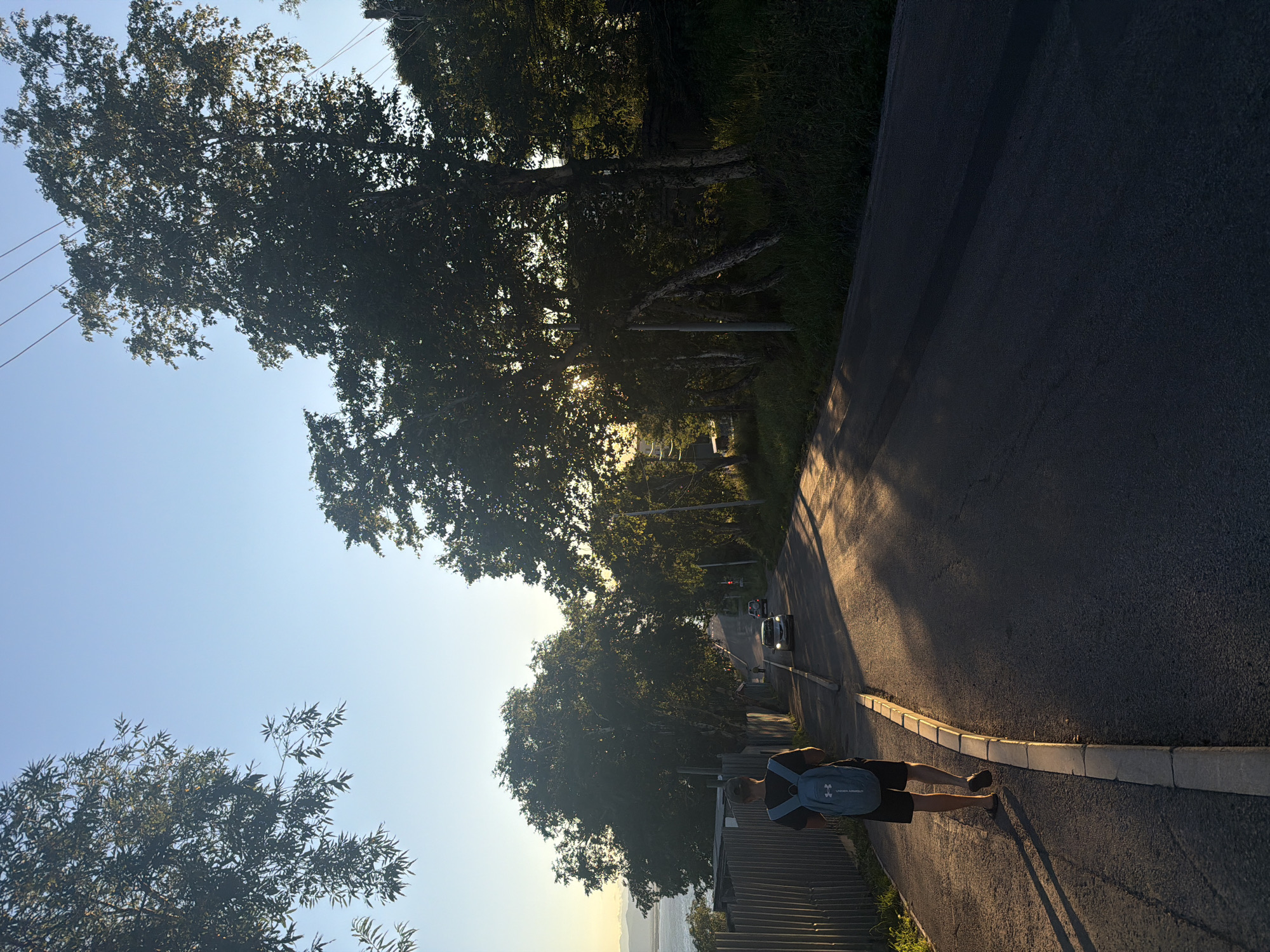A young person with a blue backpack walks down a paved road on an incline in Petropavlovsk-Kamchatsky, Russia. The person is wearing black shorts and a dark t-shirt, with sandals on their feet. The sun is setting, casting long shadows across the road and illuminating the trees on the right side of the image. The sky is clear and blue, with a hint of orange near the horizon. In the distance, there are a few cars on the road, and a body of water is visible to the left, along with a fence. The overall scene depicts a peaceful evening walk in a somewhat rural or suburban area of the city.