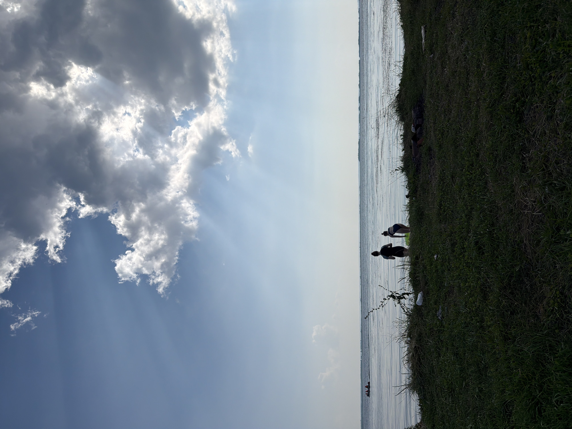 The image depicts a serene outdoor scene near the city of Krasny Bor, Russia. The foreground is dominated by a grassy embankment leading down to a large body of water, likely a river or lake. Two individuals, silhouetted against the bright sky and water, are standing near the water's edge. One appears to be wearing dark athletic attire, while the other is in swimwear. In the distance, on the water, a small boat with at least three occupants is visible. The sky is a striking feature, with dramatic clouds filtering sunlight, casting rays that create a luminous effect. The overall atmosphere is tranquil, suggesting a peaceful day by the water.
