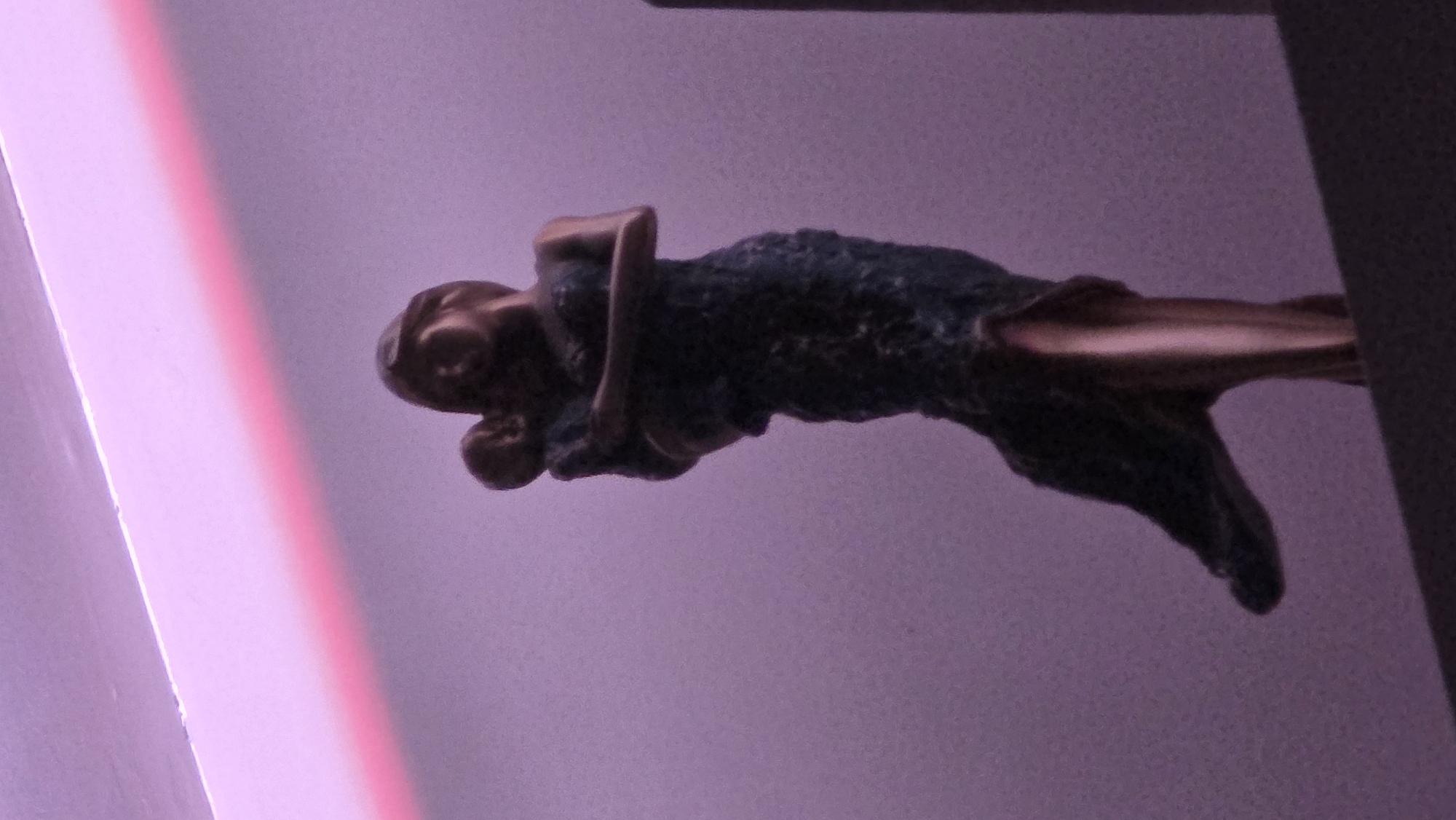 The image shows a bronze or brass sculpture of a female figure hugging a child, seemingly attached to a structure. The sculpture appears to be a gargoyle, potentially acting as a water spout, given the tail-like appendage extending outwards. The setting is indoors, with a purple-toned wall serving as the background. The context of the location within Sofia, Bulgaria, cannot be determined from the given image.