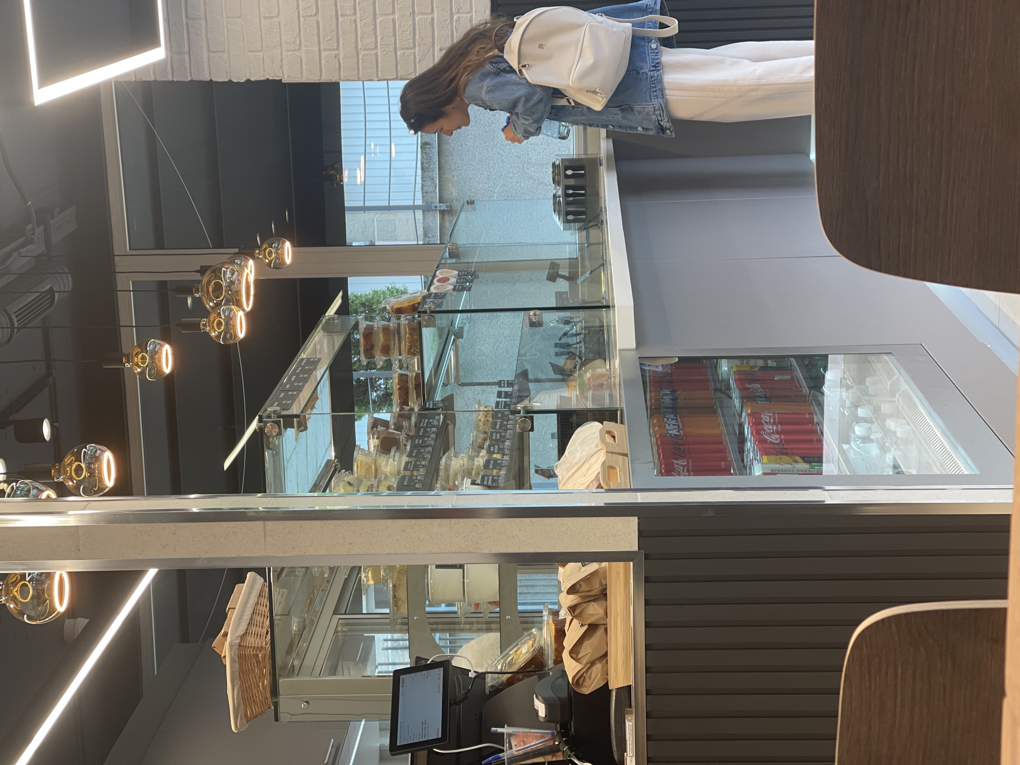 The image shows the interior of a commercial establishment, possibly a bakery or café, in Sofia, Bulgaria. A woman with long brown hair, wearing a denim jacket and white pants and carrying a white backpack, stands facing a glass display case. Inside the case are various food items such as pastries and sandwiches. Behind the display case is a cooler stocked with beverages including Coca-Cola and bottled water. Below the display is a counter with a point-of-sale system, including a monitor and other payment-related equipment. Additional baked goods are displayed above the counter. The lighting consists of modern hanging globe lights and rectangular overhead lights. The walls are a mix of white brick and dark gray horizontal panels. A chair is visible in the foreground.