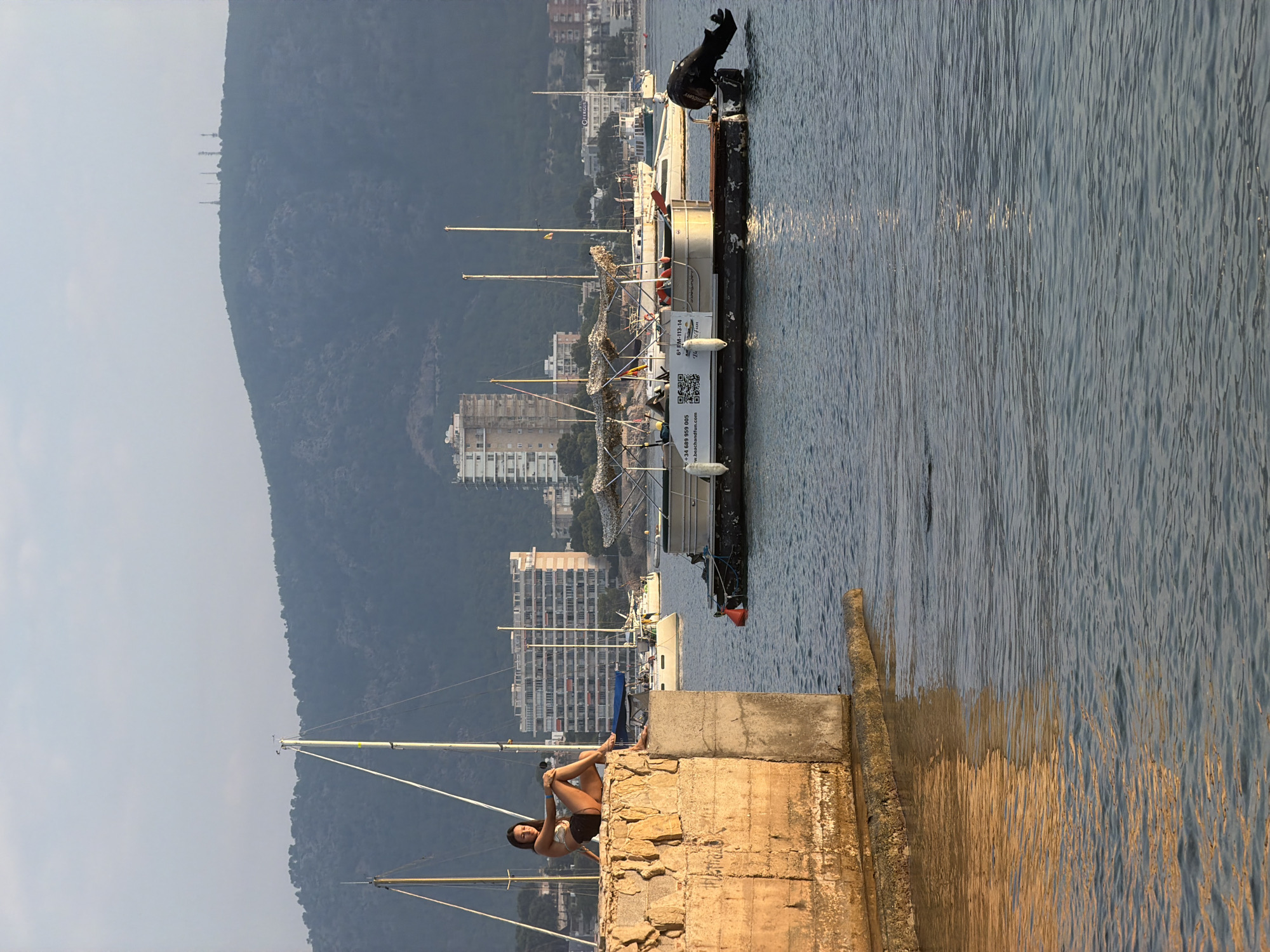 A young woman is sitting on a stone wall in the foreground of the image. She is wearing a bikini and has her legs crossed. The wall is made of rough-hewn stone and is weathered. Behind her, there is a body of water with several boats. In the background, there are buildings and a hill with trees. The lighting suggests it is either sunrise or sunset. The scene is likely Calvià, Spain, given the architectural style of the buildings and the type of boats.