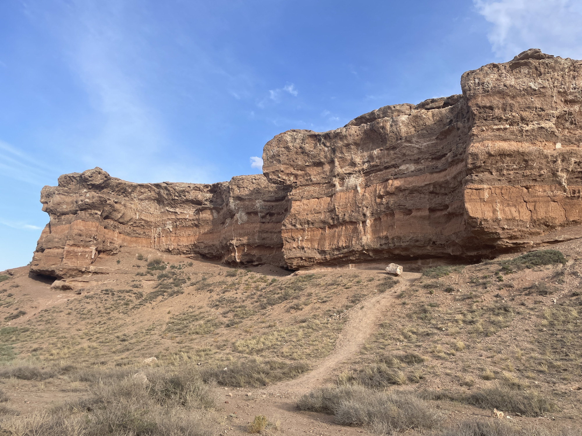 The image shows a wide, arid landscape under a bright blue sky with sparse white clouds. The dominant feature is a large, stratified rock formation, likely sandstone, with layers of reddish-brown and orange hues. The formation stretches across the middle ground and background of the image, featuring overhangs and erosional features. The foreground consists of a dusty, dry terrain covered with low-lying, sparse, dry grass and bushes. A dirt path winds its way up the gentle slope towards the rock formation. In the middle ground, near the base of the rock formation, a large, light-colored boulder is visible. There are no people or animals in the frame. The scene is indicative of a natural, undeveloped environment. The location is identified as being in the city of Tamerlik, Kazakhstan, suggesting a desert or semi-desert climate.