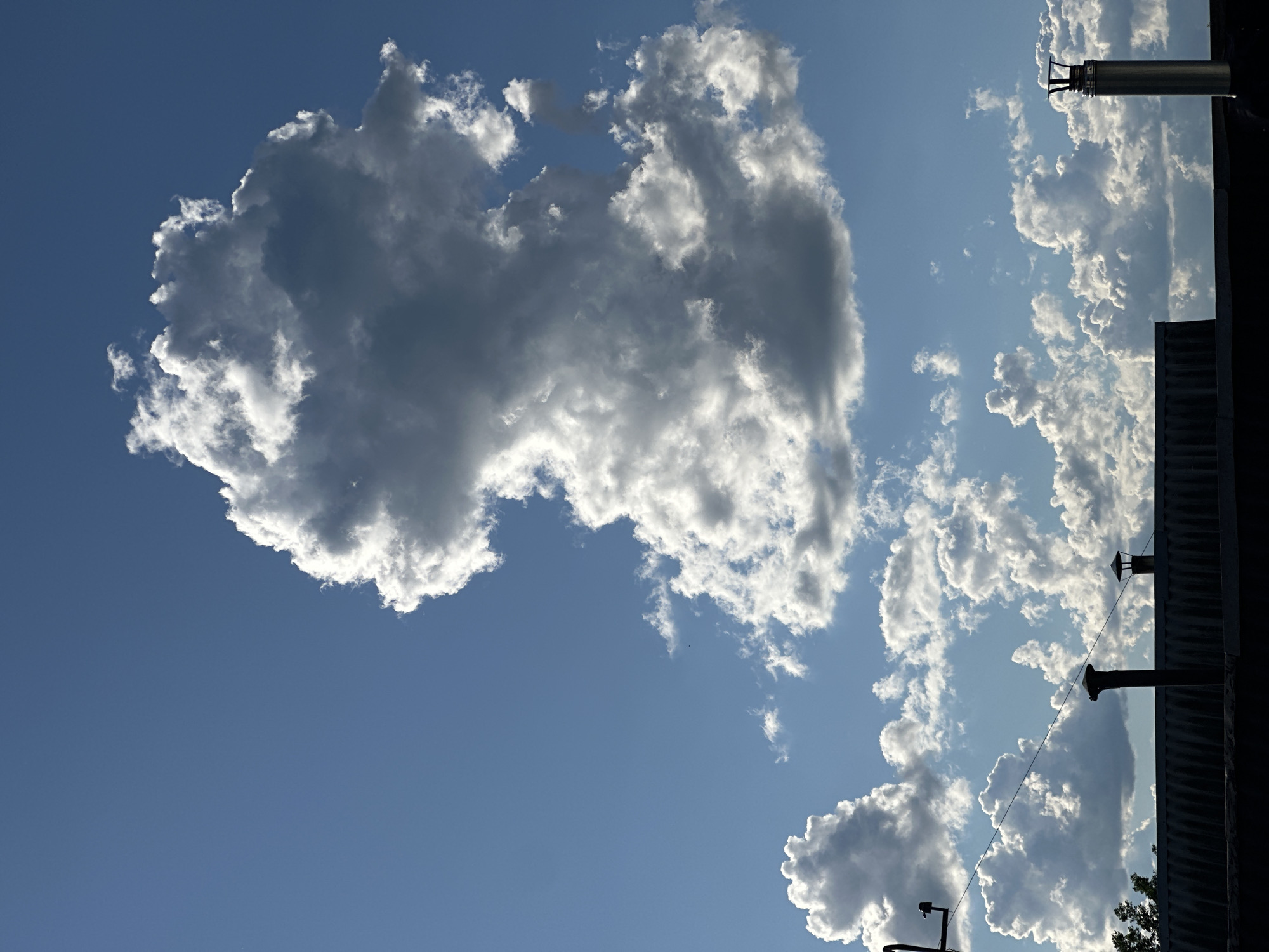 The image shows a view of the sky in Chaikovsky, Russia. A large, cumulus cloud dominates the left side of the frame, its white edges reflecting sunlight against a backdrop of blue sky. Other, smaller cumulus clouds are scattered to the right, partially obscured by the silhouette of a building. The building's edge is visible in the upper right and bottom right of the image, featuring ventilation pipes extending upward. The image is taken at a slight angle, with the top of the building seemingly tilted. The scene appears to be captured during daylight. There are no people visible in the image.