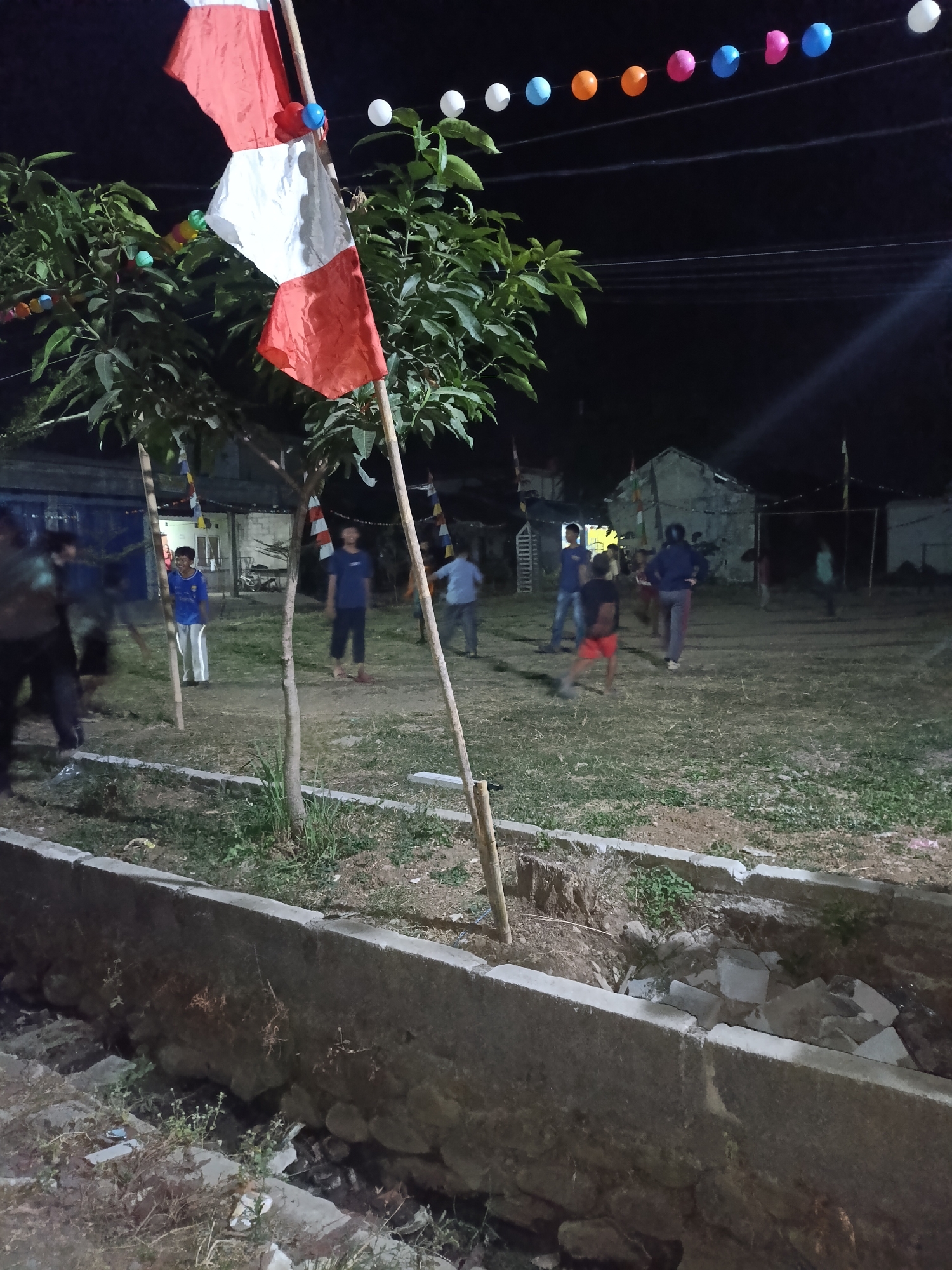 In Cariu, Indonesia, this nighttime photograph captures a scene with several young people engaged in an outdoor activity. In the foreground, a concrete channel filled with debris is visible. Beyond this, a grassy area appears to be the site of a gathering. Two trees stand prominently, one bearing the Indonesian flag, a red and white bicolor flag, and decorative colorful ball-shaped lights strung between poles and trees. Several individuals are dispersed across the field, some appearing to be playing or interacting. In the background, a few simple structures, possibly houses, are dimly lit, suggesting a residential or community area. The overall atmosphere is casual and communal, illuminated by artificial light sources.