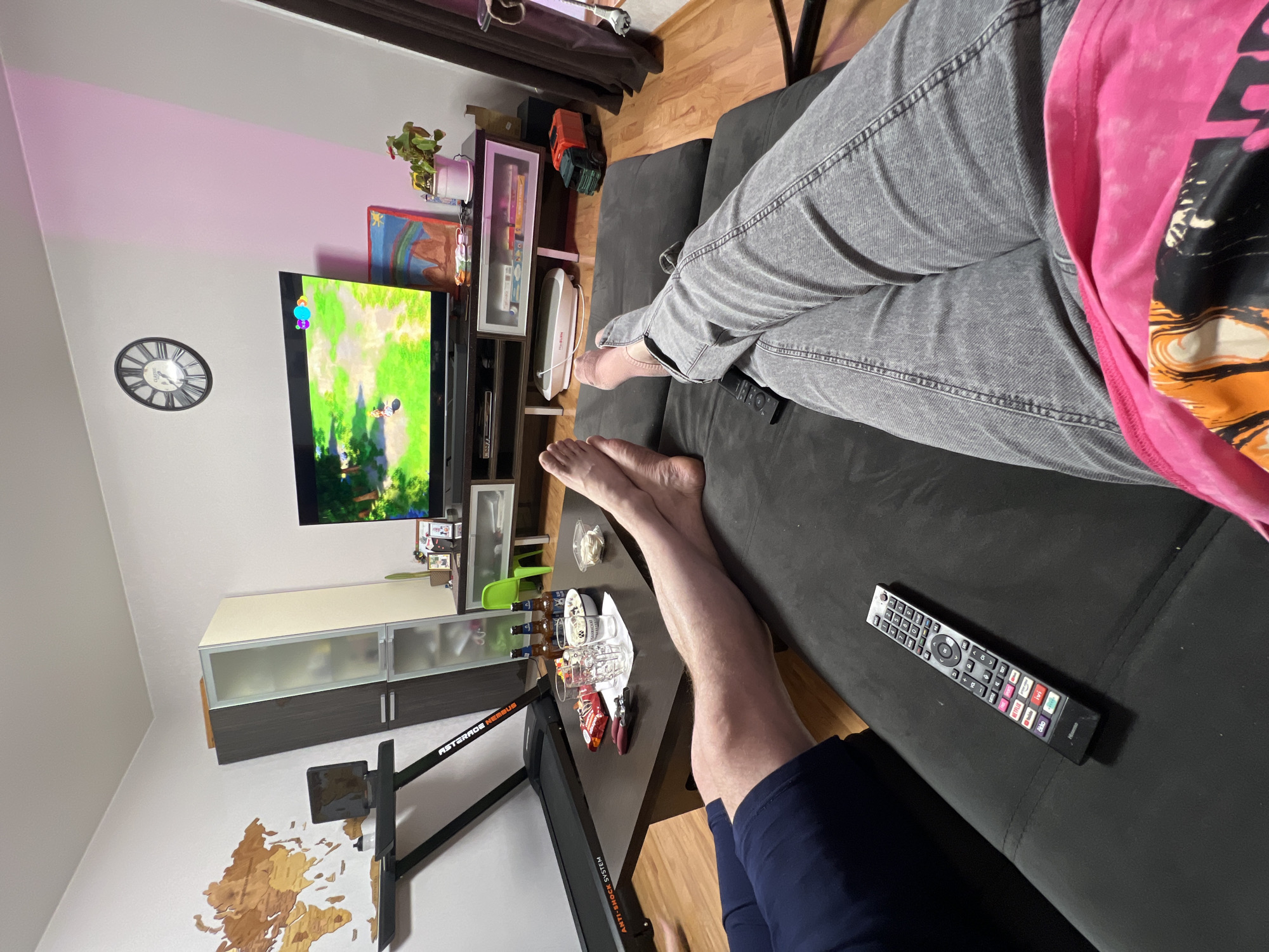 This image captures a casual indoor scene, likely a living room in Perm, Russia, from a high-angle, first-person perspective. Two individuals are present, relaxing.
The person taking the photo has their bare feet and lower legs visible, wearing dark blue shorts, with their legs crossed and propped on a dark wooden coffee table. Opposite them, another person is lounging on a dark grey sofa or chaise lounge, with their bare feet visible and legs crossed. This individual is wearing light grey jeans and a pink t-shirt with a graphic design.
The primary activity appears to be watching television. A flat-screen TV displays animated content, resembling a cartoon or video game. A black "Hisense" brand remote control, featuring dedicated buttons for "Netflix," "YouTube," and "Prime Video," rests on the sofa.
The coffee table in the foreground holds several items, including three empty beer bottles, two empty beer glasses, a small white bowl, and a red packet of crisps or snacks. The room's decor includes a round wall clock with Roman numerals and the text "CLASSIC," a wooden world map decoration, a potted plant, and framed pictures. A pink/purple light casts an ambient glow on the wall above the television. Other notable objects include a two-toned TV cabinet, a small white appliance on the stand, and part of a piece of exercise equipment (possibly a treadmill) with "ASTERADE NIMBUS" and "ANTI-SHOCK SYSTEM" visible on its frame. The overall impression is one of a relaxed, casual evening at home.