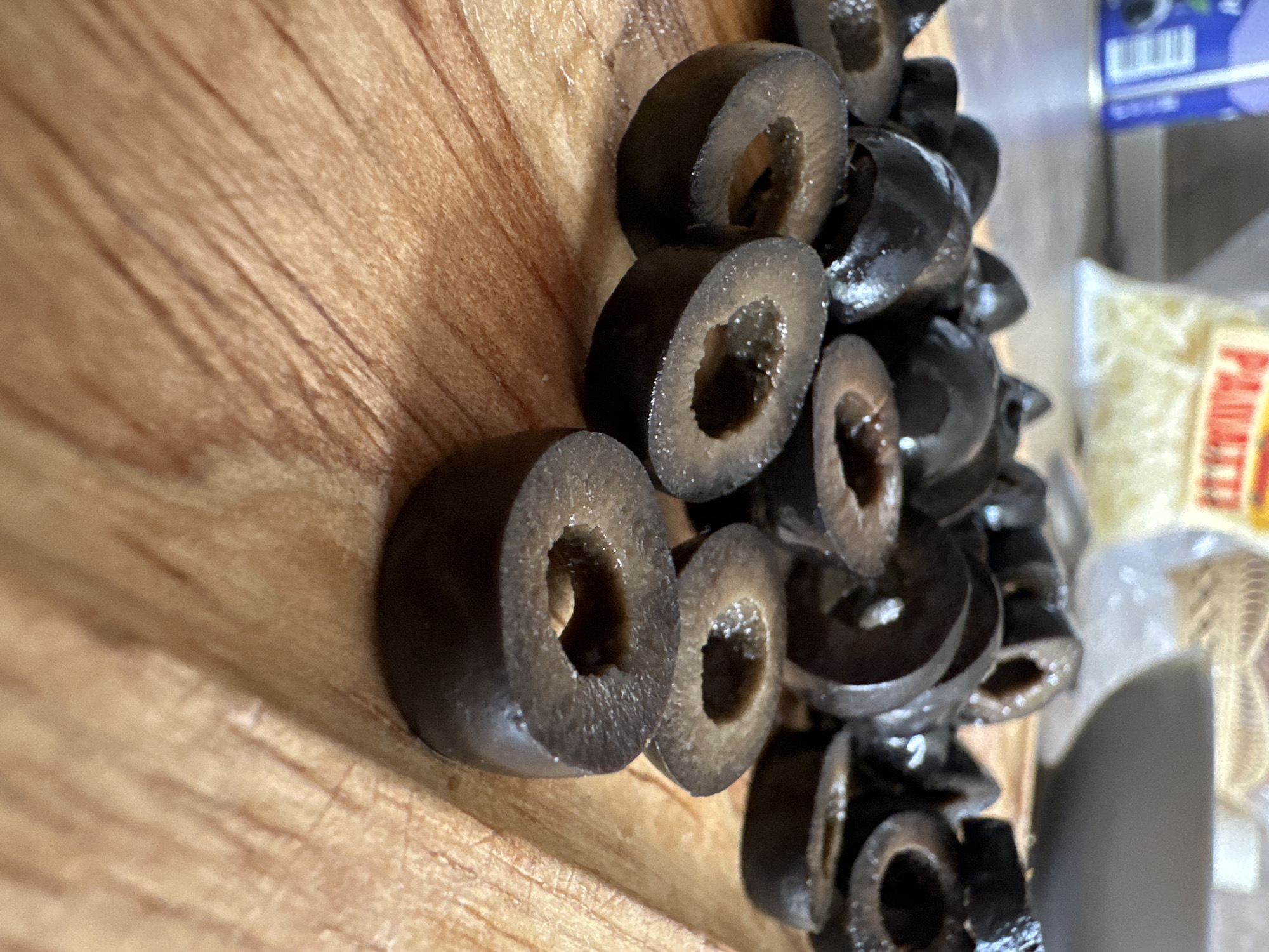 The image presents a close-up, slightly overhead view of sliced black olives resting on a wooden cutting board. The olives are dark, ranging from deep brown to black, and many exhibit a glistening surface, suggesting moisture. Their distinct hollow centers are clearly visible from their cross-sections. The wooden surface displays prominent grain patterns.
The olives are primarily concentrated on the right side of the frame, with some scattered towards the center and lower left. In the background, to the right, several other items are partially visible. A blue package, likely an olive container, shows a barcode and the word "OLIVES." Below this, a transparent plastic bag contains shredded, light-colored material, strongly resembling grated cheese, with part of a label reading "PARMIG..." (suggesting Parmesan) visible. A blurred gray object, possibly a kitchen item, is also present at the bottom right.
The scene is set indoors, most probably in a kitchen, indicating food preparation. No people are visible. The lighting suggests it is daytime. While the image itself does not contain specific visual cues to confirm the location, the provided context places this scene in Stavropol, Russia.