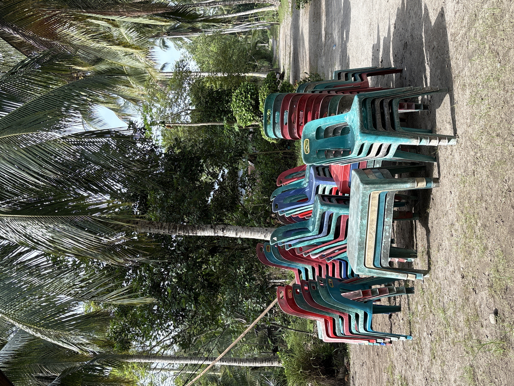 In the village of Beunyot, Indonesia, an outdoor scene features stacks of colorful plastic chairs, primarily red, teal, and blue, arranged in front of lush green foliage and tall palm trees. A dirt path leads into the distance, lined with more palm trees. One stack of teal chairs is prominent in the foreground, with a teal and white plastic table to its left. The ground is covered in dry dirt and sparse patches of grass, with a small rock visible. The lighting suggests a bright, sunny day.
