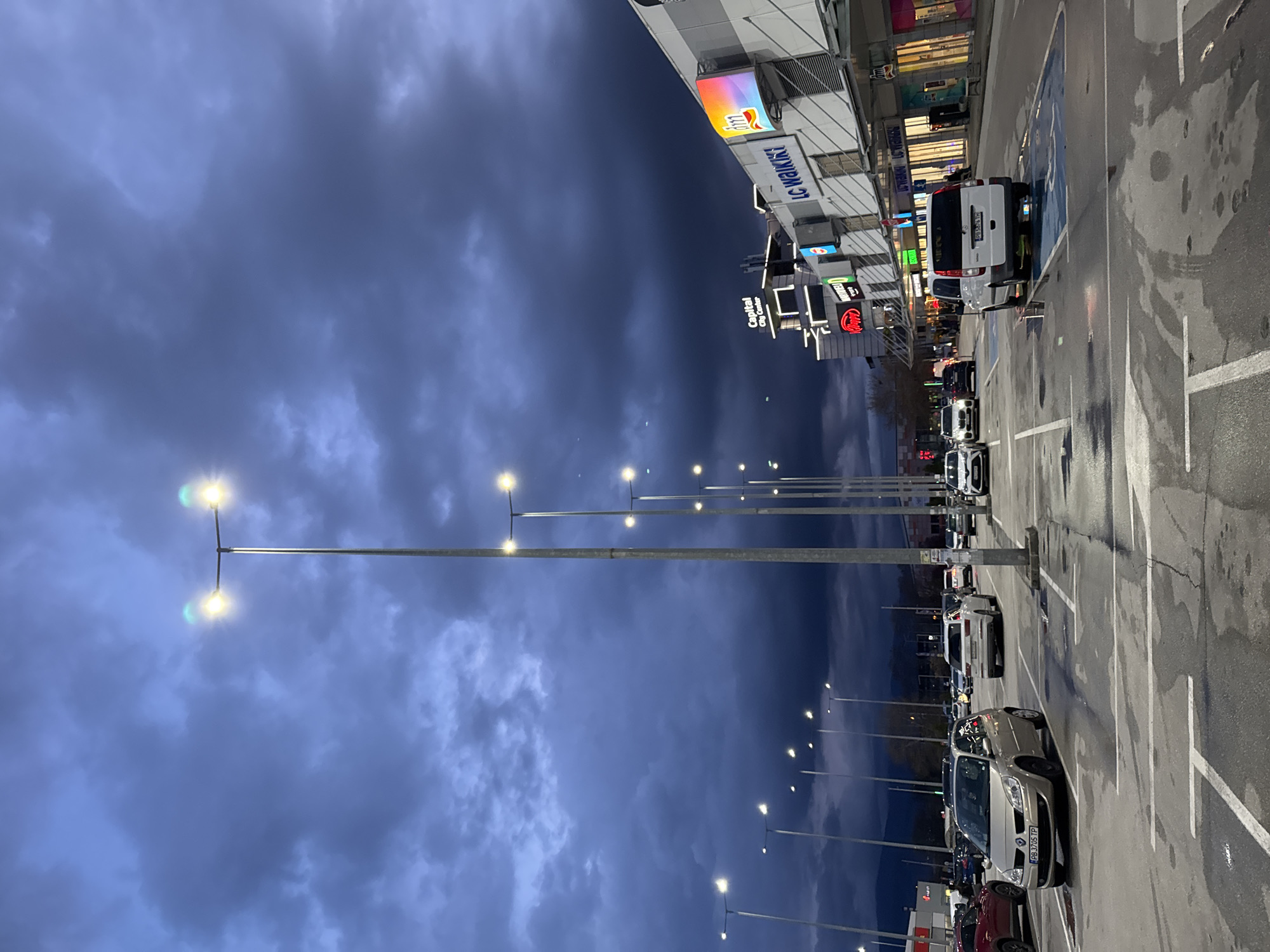 The image depicts an outdoor parking lot, seemingly at dusk or night, outside a shopping center in Plovdiv, Bulgaria. The sky is overcast with dark, dramatic clouds. Rows of streetlights illuminate the parking area. Cars are parked in marked spaces. The shopping center has various store signs visible, including "LC Waikiki," "Capital City Center," and a Coca-Cola sign. A white van is parked in a handicapped space. The pavement shows signs of recent rain or dampness. No people are clearly visible, but there are hints of people near the shopfront.