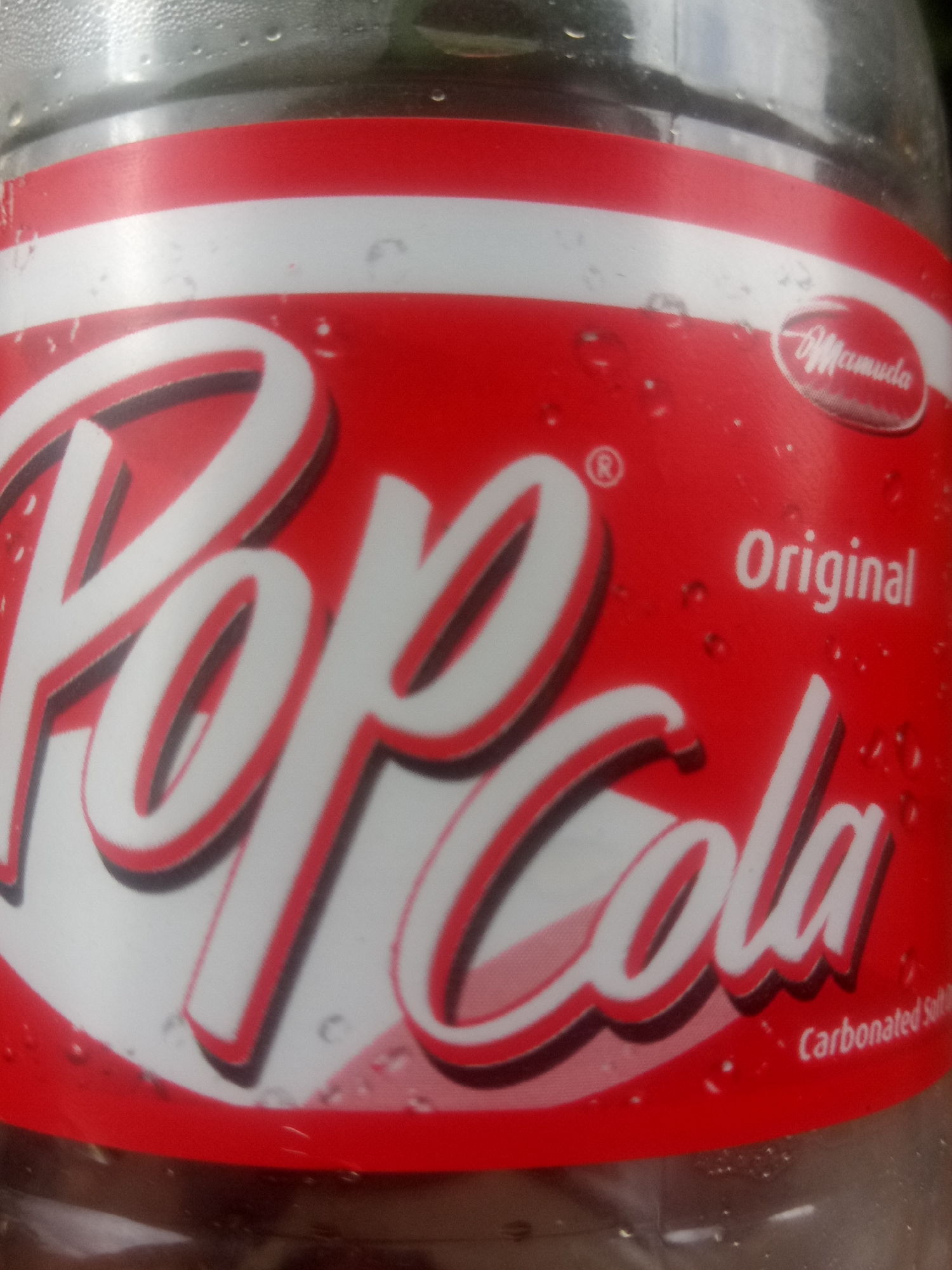 The image shows a close-up of a plastic bottle of "Pop Cola" flavored carbonated drink. The label is predominantly red with white and black lettering. The large, stylized text "Pop Cola" is the main feature, with "Pop" in a thicker font and "Cola" in a script font. Above "Cola," the word "Original" is written in smaller white text. To the upper right of the "Cola" text, there is a logo that reads "Mamuda" in a script font within an oval. Below the "Cola" text, smaller white text indicates "Carbonated Soft Drink". The bottle appears to be cool, with condensation droplets visible on its surface, suggesting it may be refrigerated or recently removed from a cold environment. The background is out of focus, but appears to be green, possibly indicating an outdoor setting or the presence of plants. The lighting suggests daytime. There are no people visible in the image.